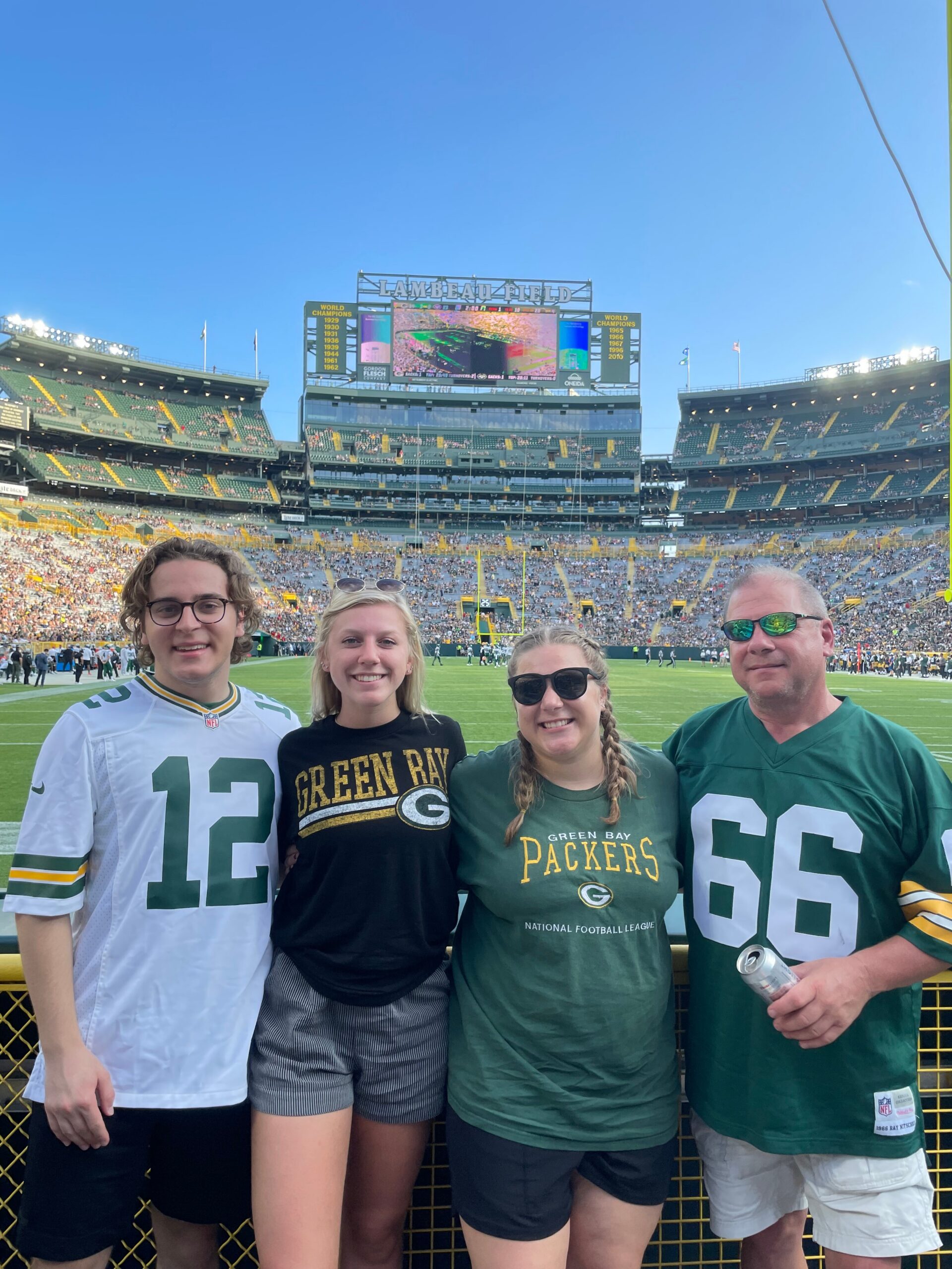 Four Packers fans posing together at Lambeau Field, wearing team jerseys and green gear with the scoreboard behind them on a sunny day.