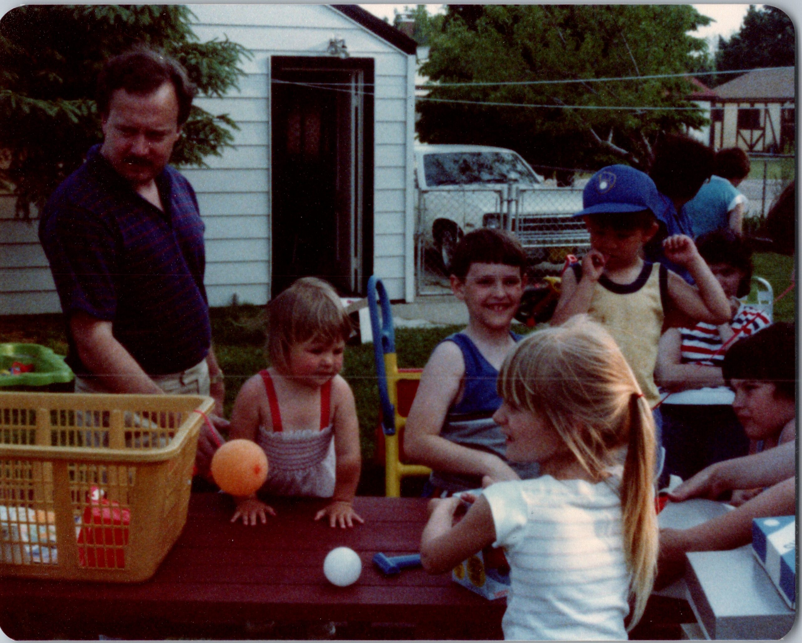 Children gathered around a table outdoors for a backyard party, with a man standing on the left and colorful toys nearby.