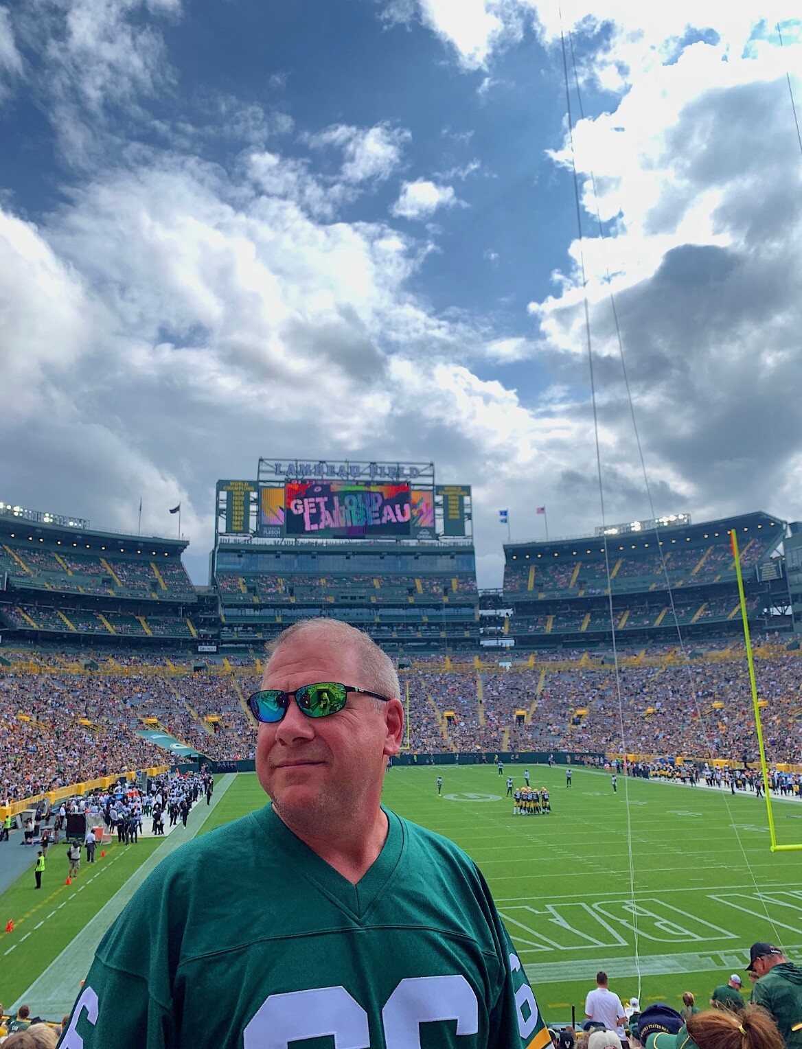 Man in green jersey and sunglasses stands in a football stadium with a large crowd and a colorful scoreboard in the background.