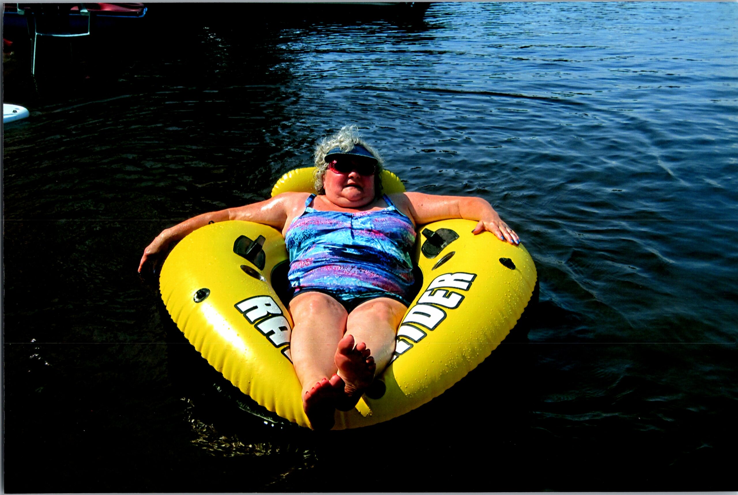 Person relaxing on a bright yellow inflatable tube on dark blue water, wearing a blue patterned swimsuit and sunglasses.