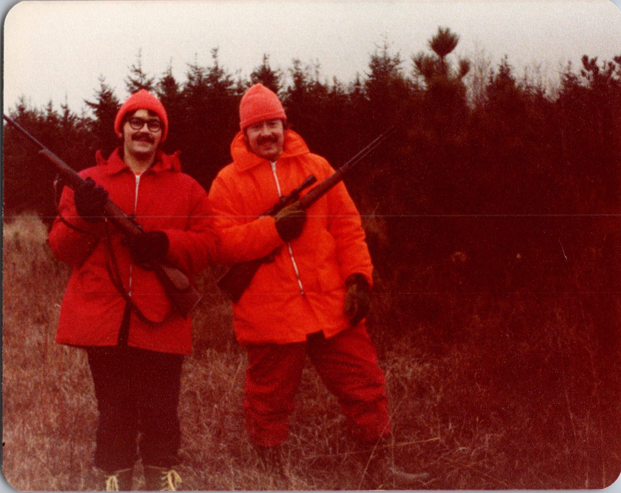 Two people in bright orange jackets stand in a forest, each holding a rifle and smiling at the camera.