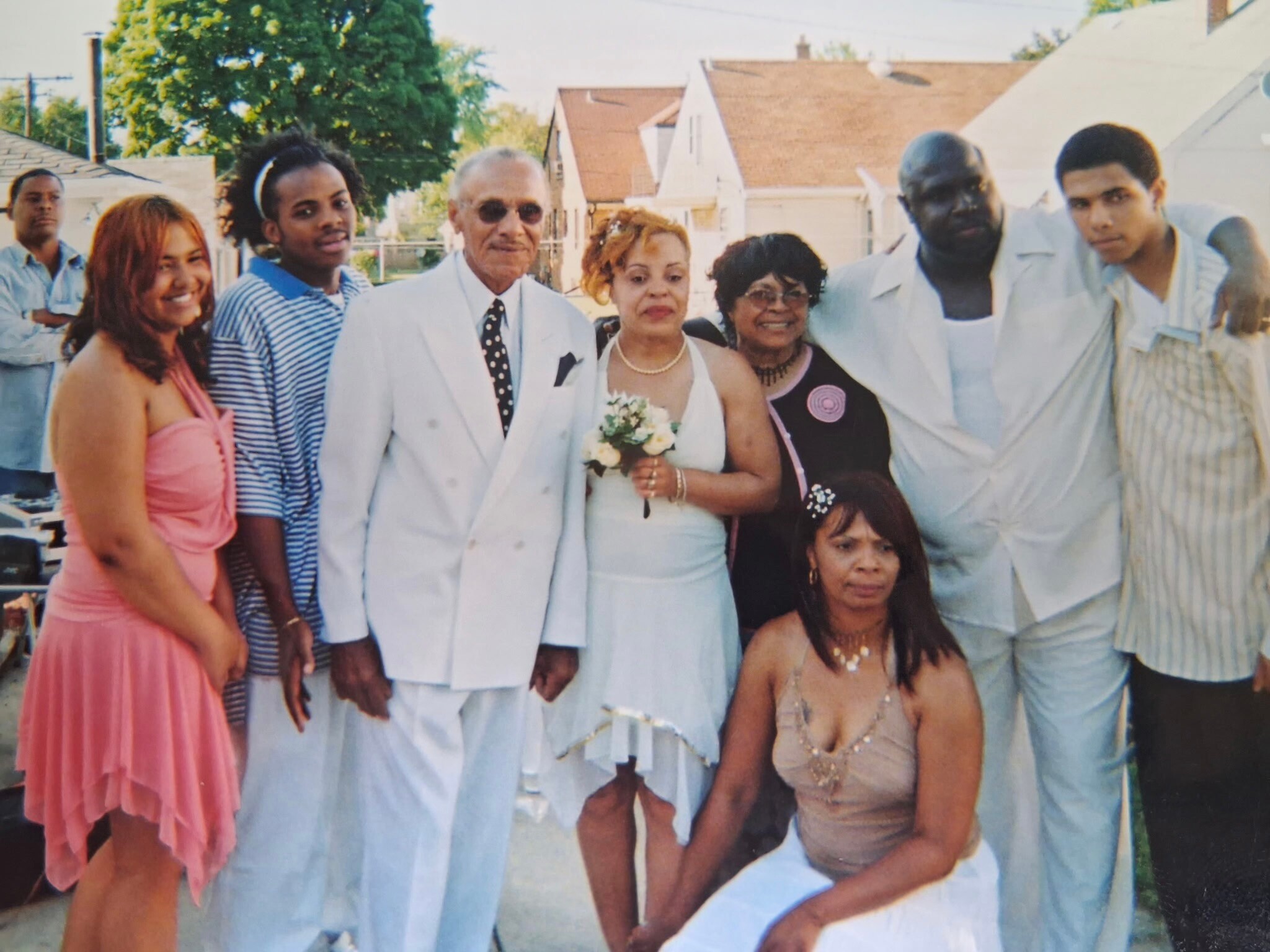 Group of diverse wedding guests posing outdoors with the bride in a white gown and bouquet in the center.