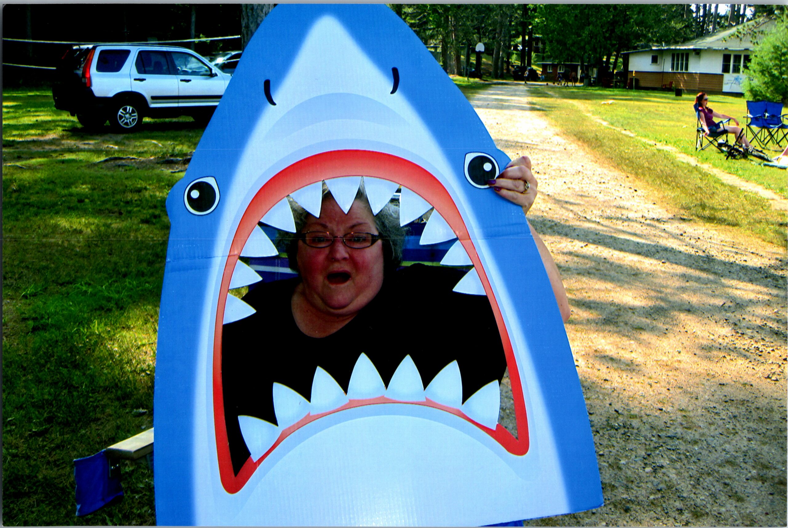 Person wearing a blue inflatable shark costume peaks out from its mouth in a park setting with grass and trees nearby.
