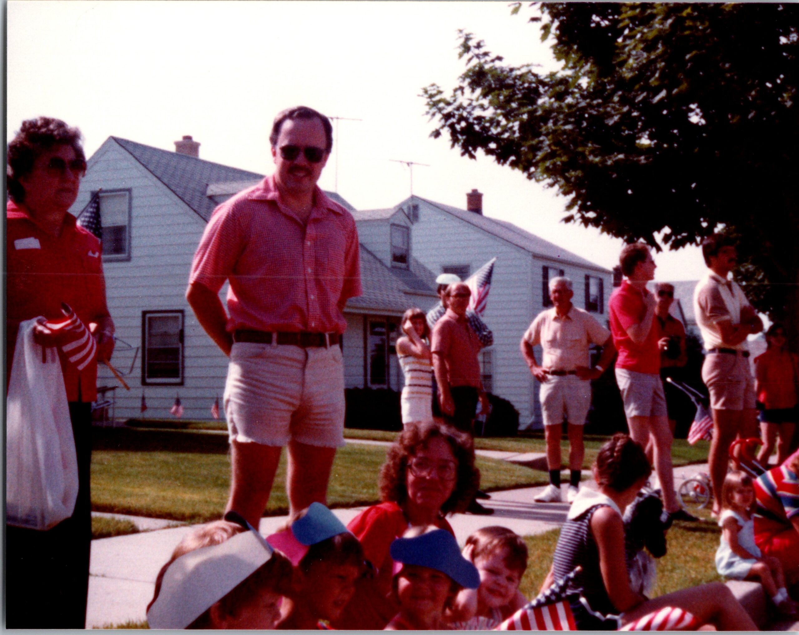 Group of people gathered on a suburban lawn and sidewalk for an outdoor event, with houses and trees in the background.