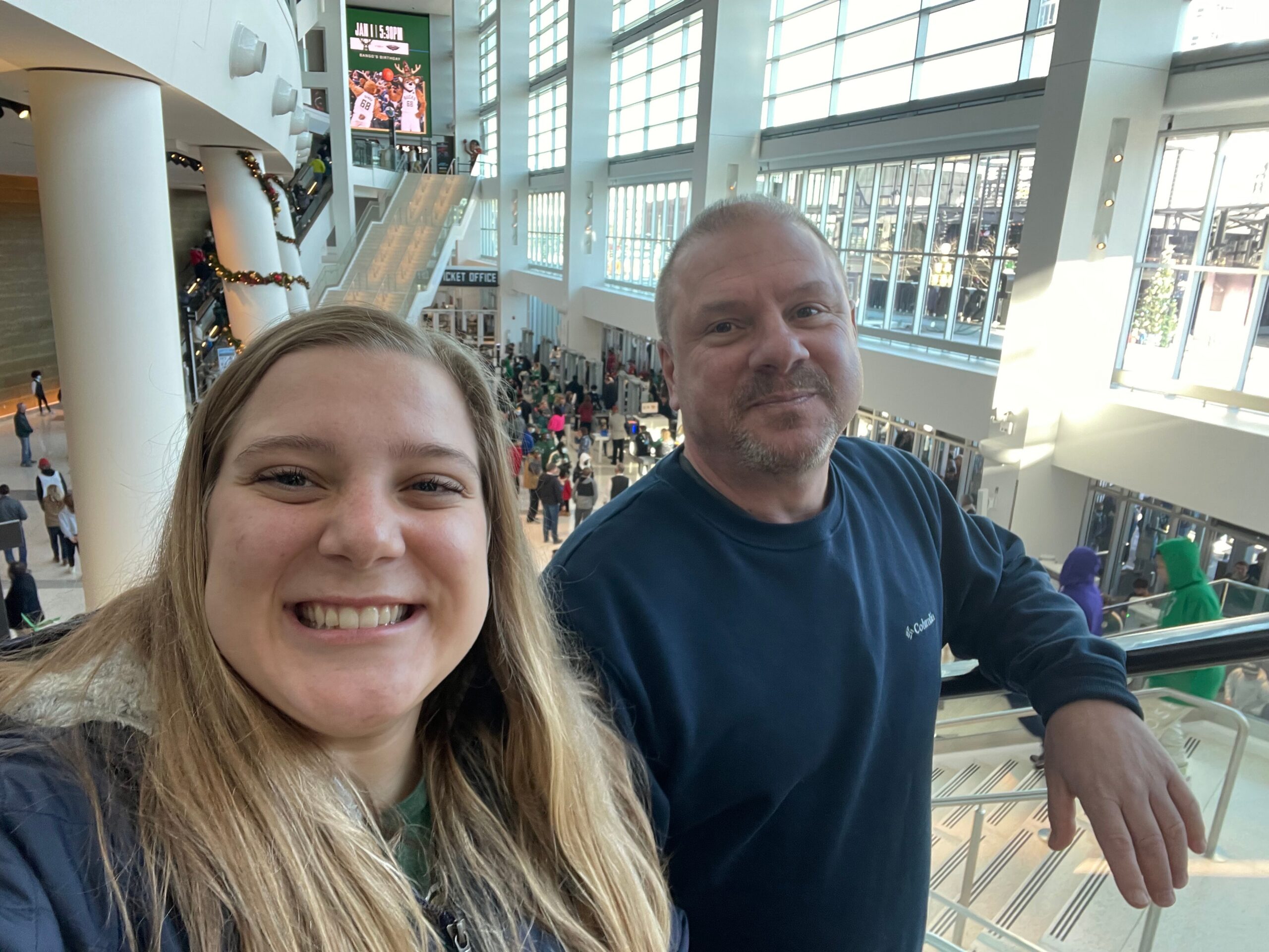 Two smiling people take a selfie in a bright, multi‑level mall atrium with escalators and shoppers in the background.