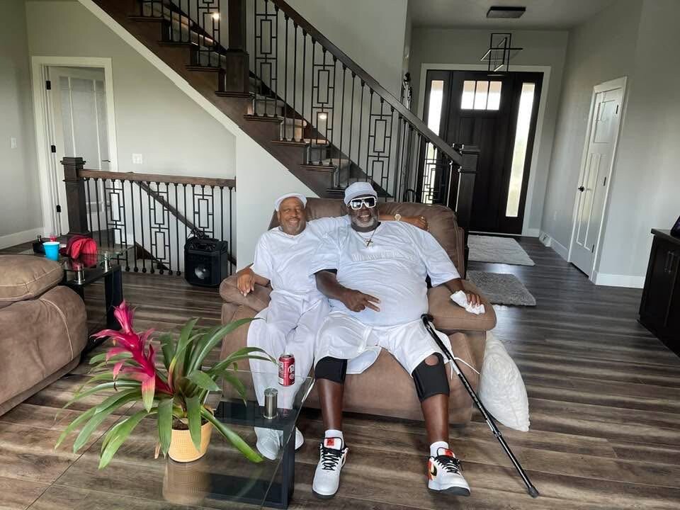 Two men sitting on a brown sofa in a bright living room, one with a cane, both smiling.