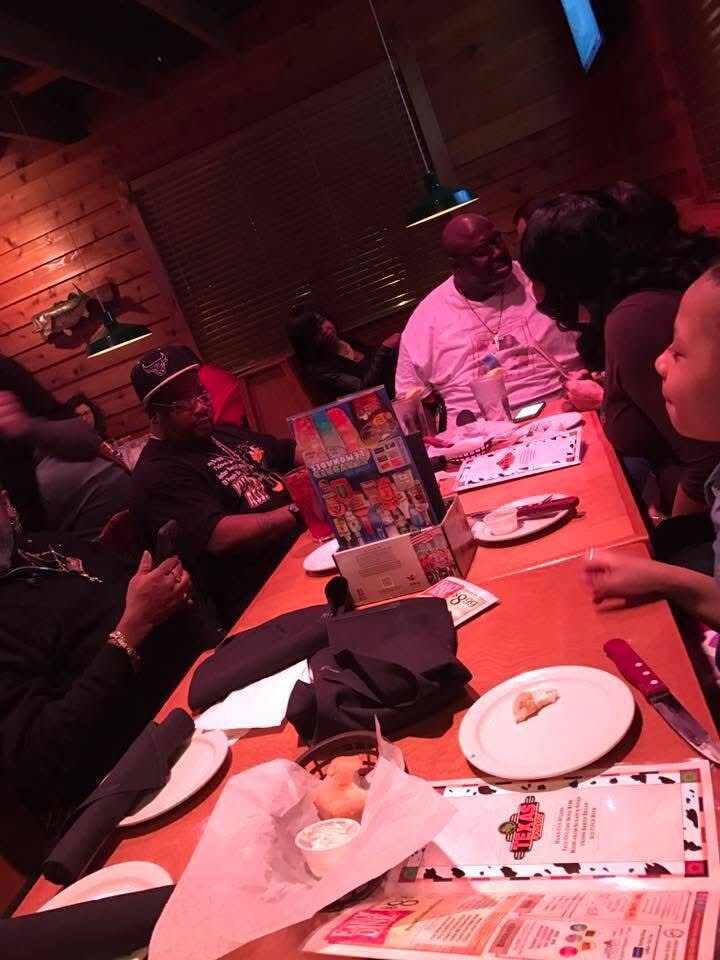 People seated around a long wooden table in a dimly lit restaurant, chatting and enjoying food and drinks.