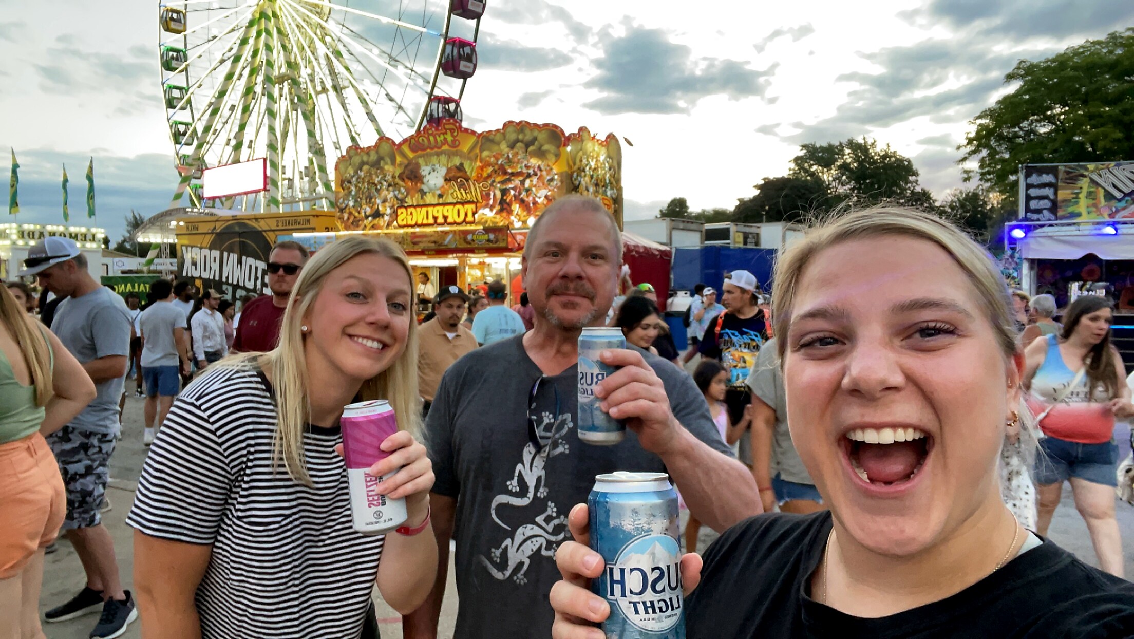 Three friends at a summer fair take a selfie, holding drinks with a large Ferris wheel and colorful rides behind them.