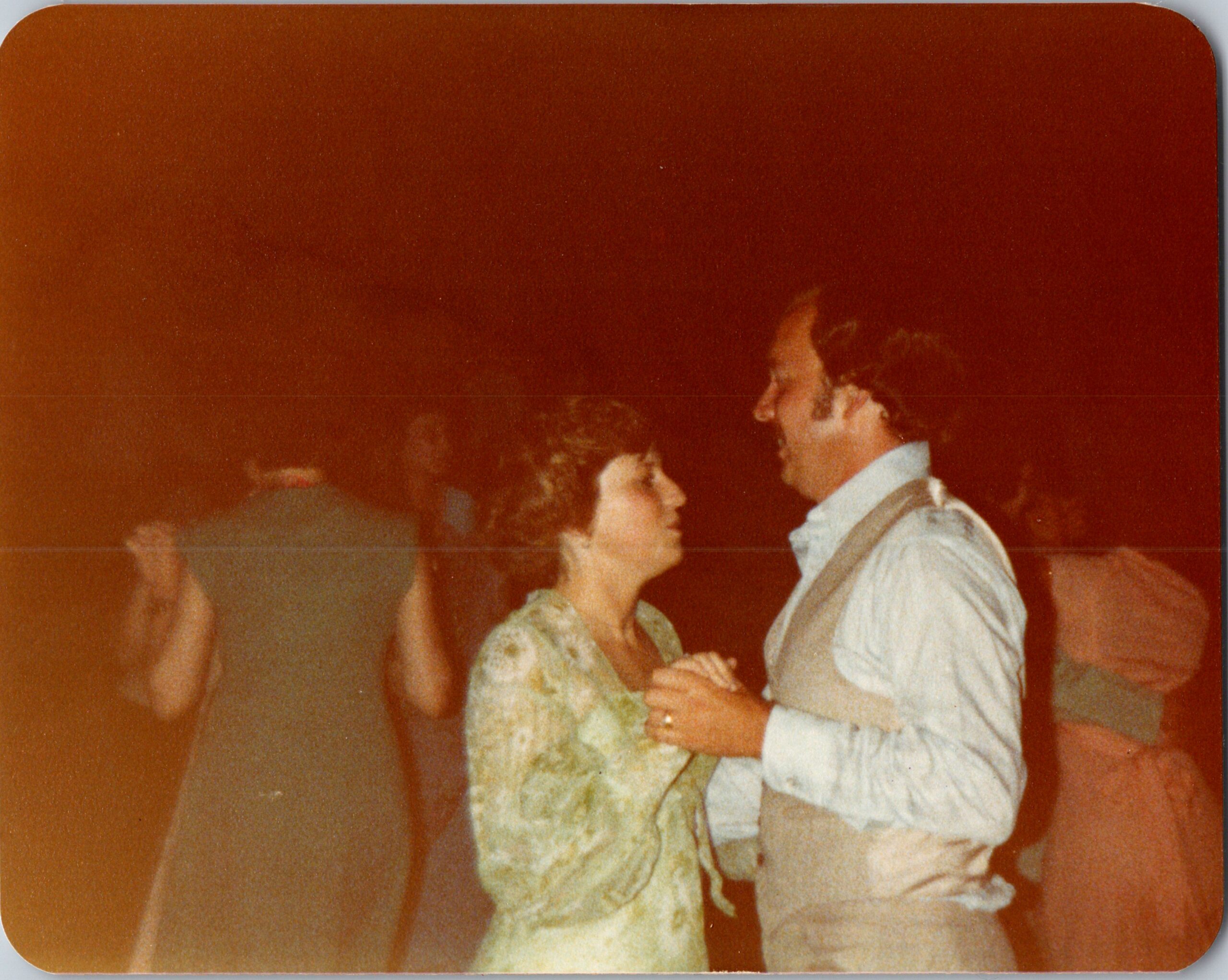 Couple dancing together in a warm, reddish-lit setting; woman in floral dress and man in light shirt with suspenders are facing each other.