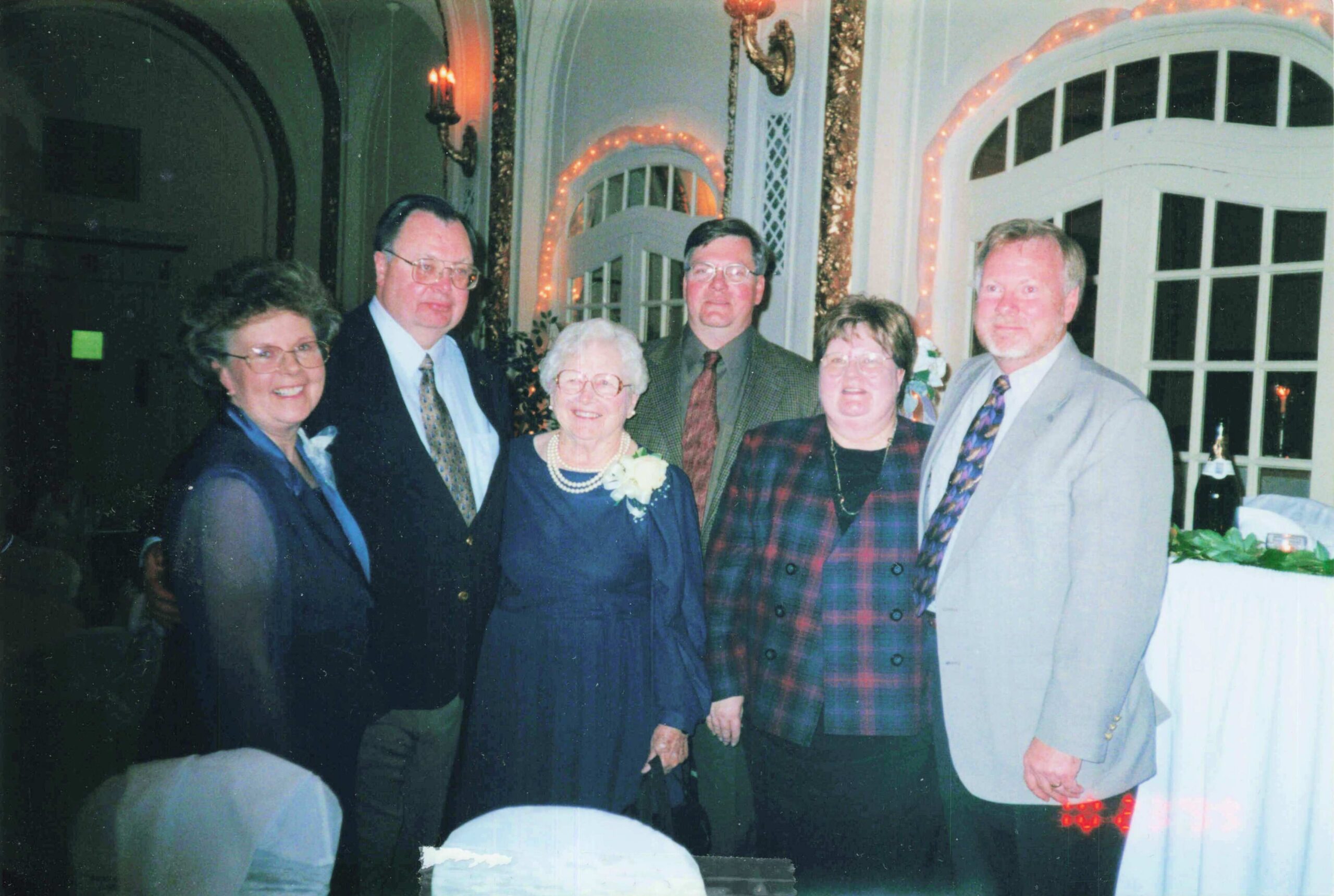 Six adults in formal attire posing for a group photo at a decorated banquet hall with string lights.