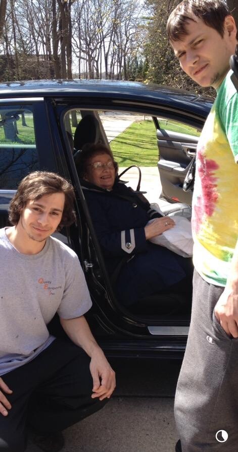 Three people pose by a black car with the rear door open: an older woman sits in the driver's seat smiling, while two younger men stand outside in daylight on a sunny day.