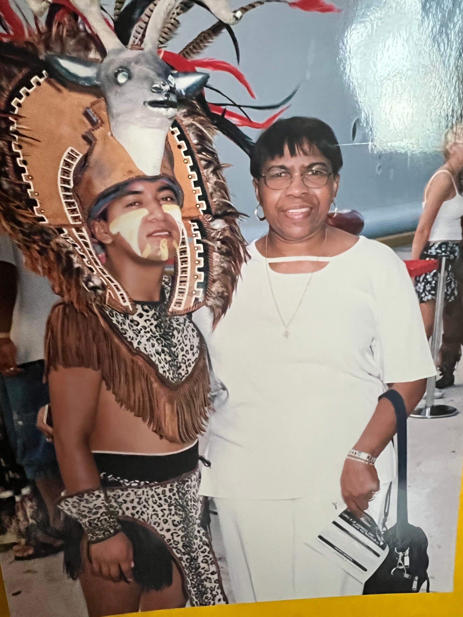 Two women posing together at a festive event; one in a white outfit and glasses, the other wearing an elaborate feathered headdress with face paint.