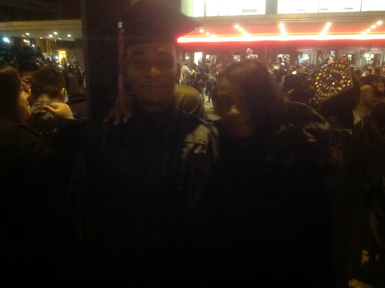 Two people stand close together in a crowded nighttime street scene with a busy storefront and neon lights in the background.