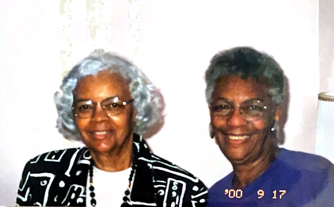 Two elderly African American women smile for a photo indoors, one wearing a black-and-white geometric jacket and glasses, the other in a purple top, standing close together.