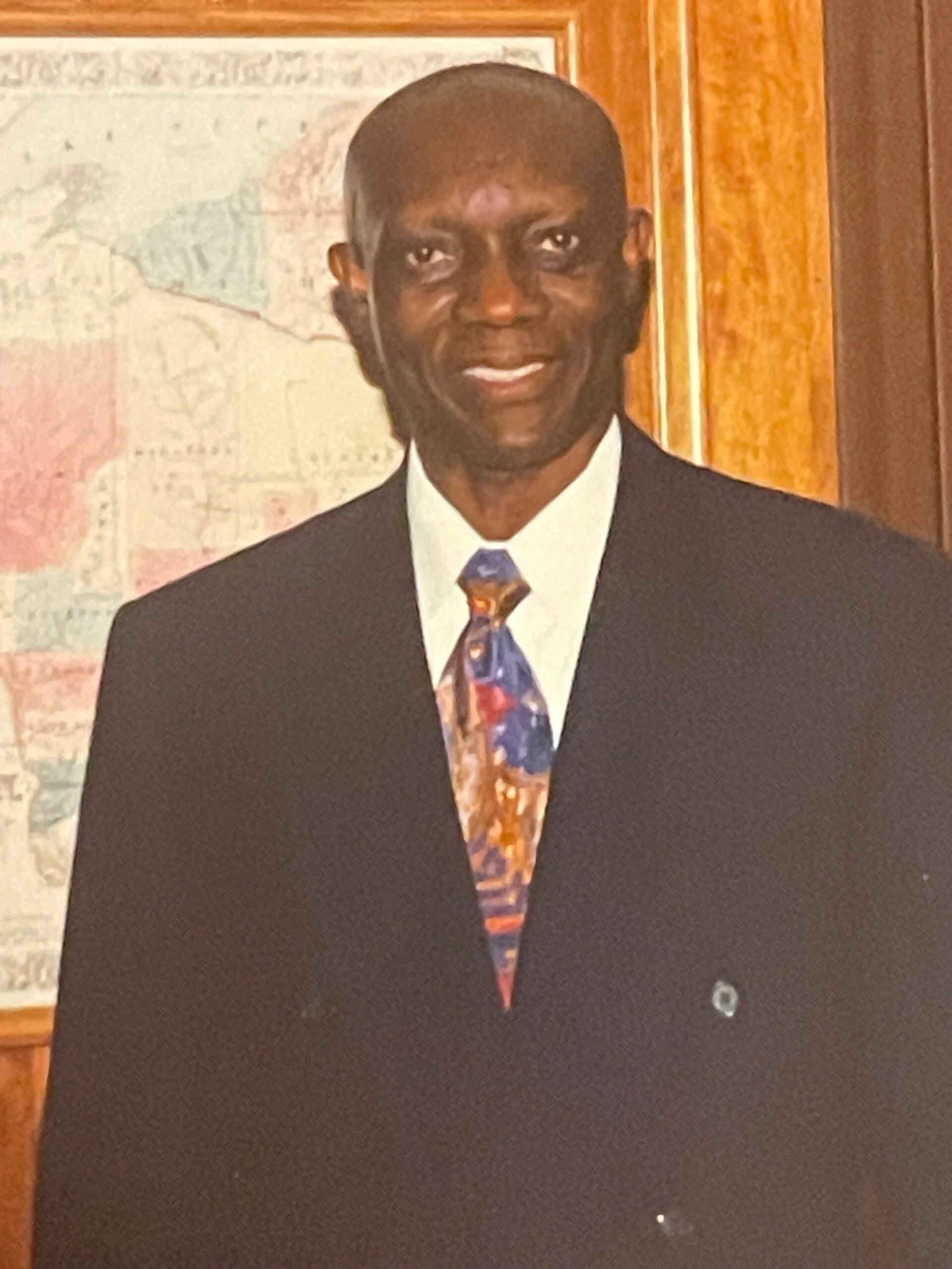 Man in a dark suit and colorful tie posing indoors, with a map on the wall in the background.