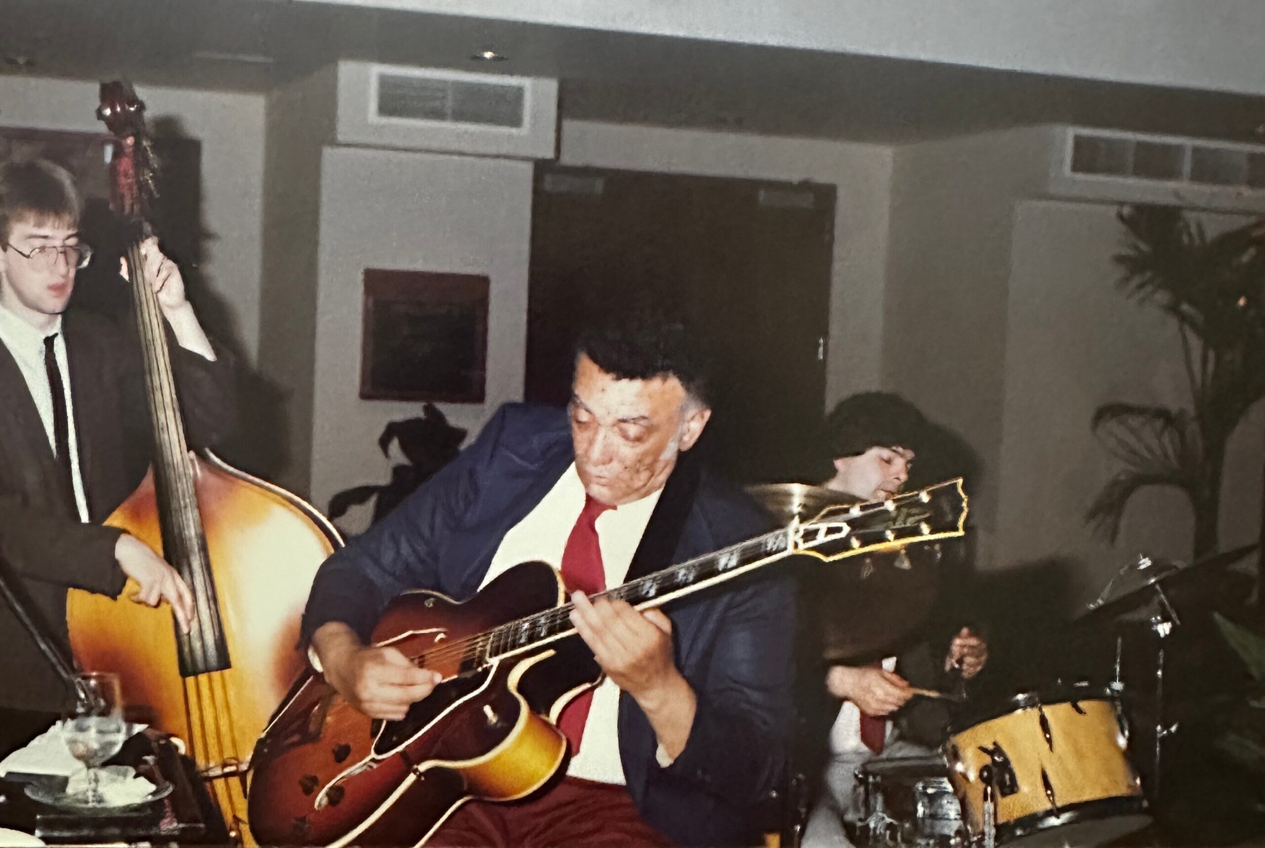 Musician in a blue blazer playing an acoustic guitar at a festive indoor gathering, other band members visible in background.