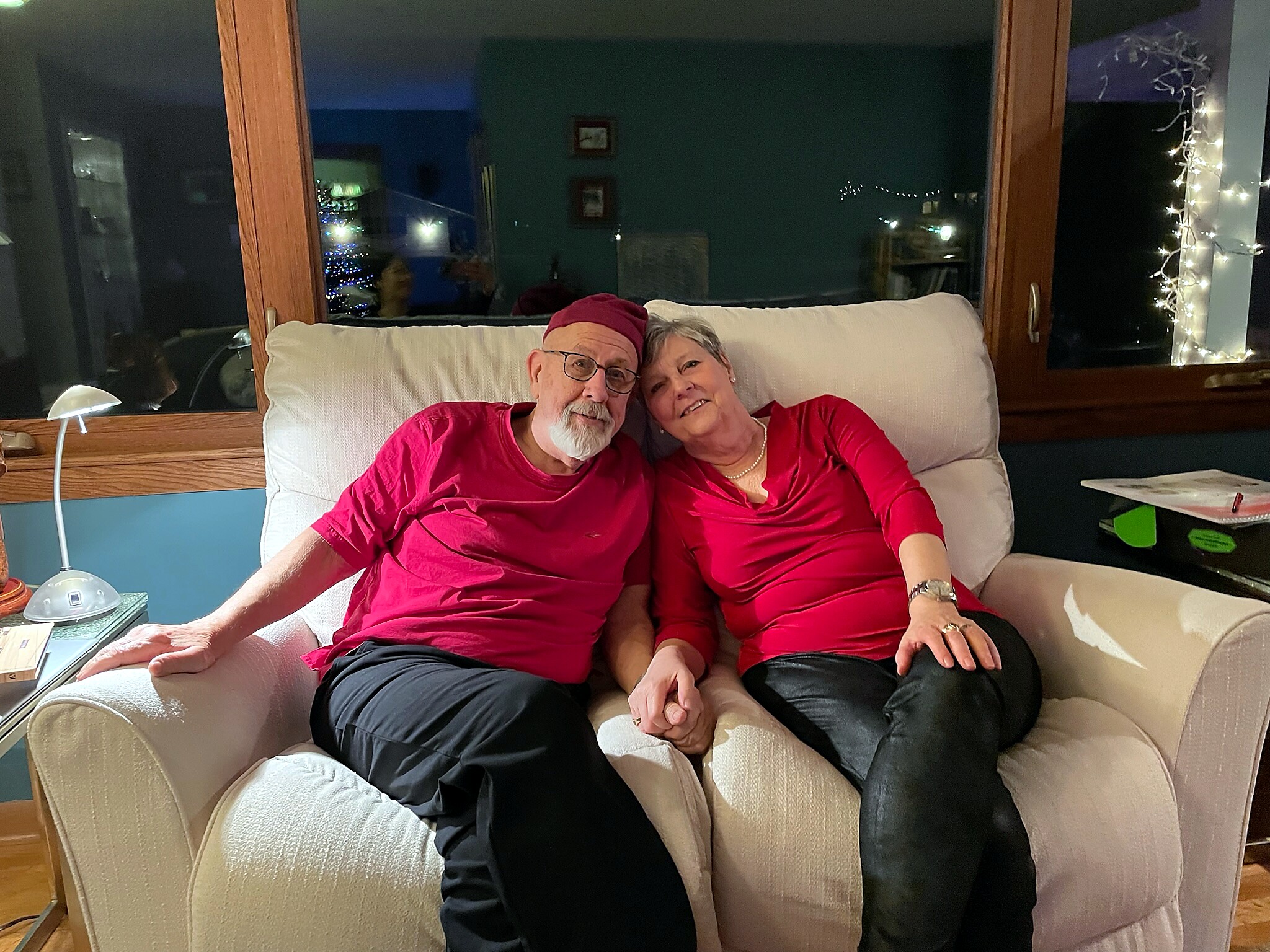 Elderly couple in red shirts sitting together on a beige sofa, smiling at the camera with fairy lights in the background.