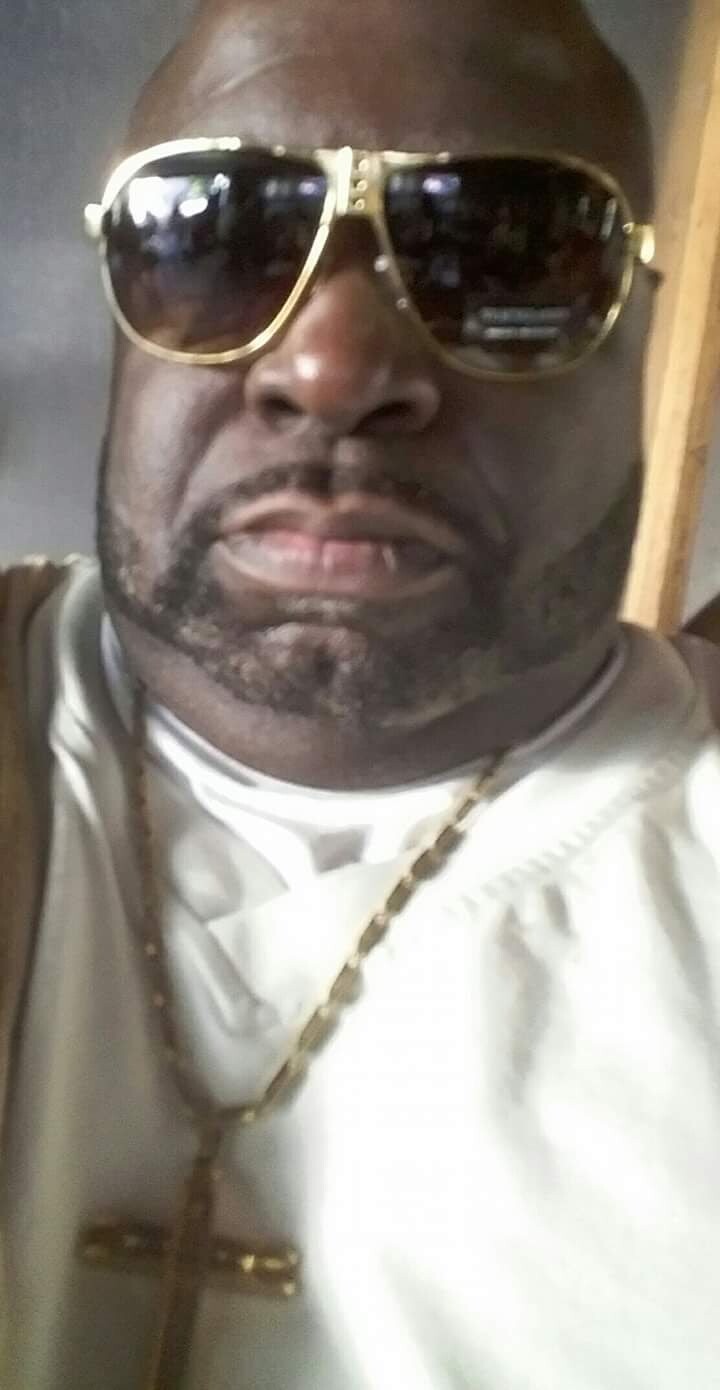 Man wearing large gold aviator sunglasses and a gold chain, in a light-colored shirt, facing the camera.
