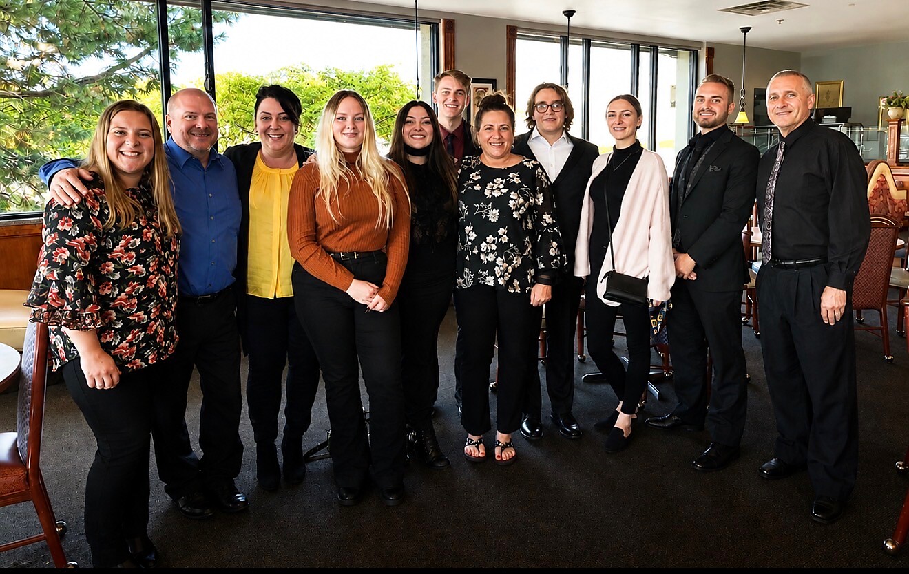 Twelve coworkers posing together in a bright dining area with large windows, dressed in smart casual clothes.