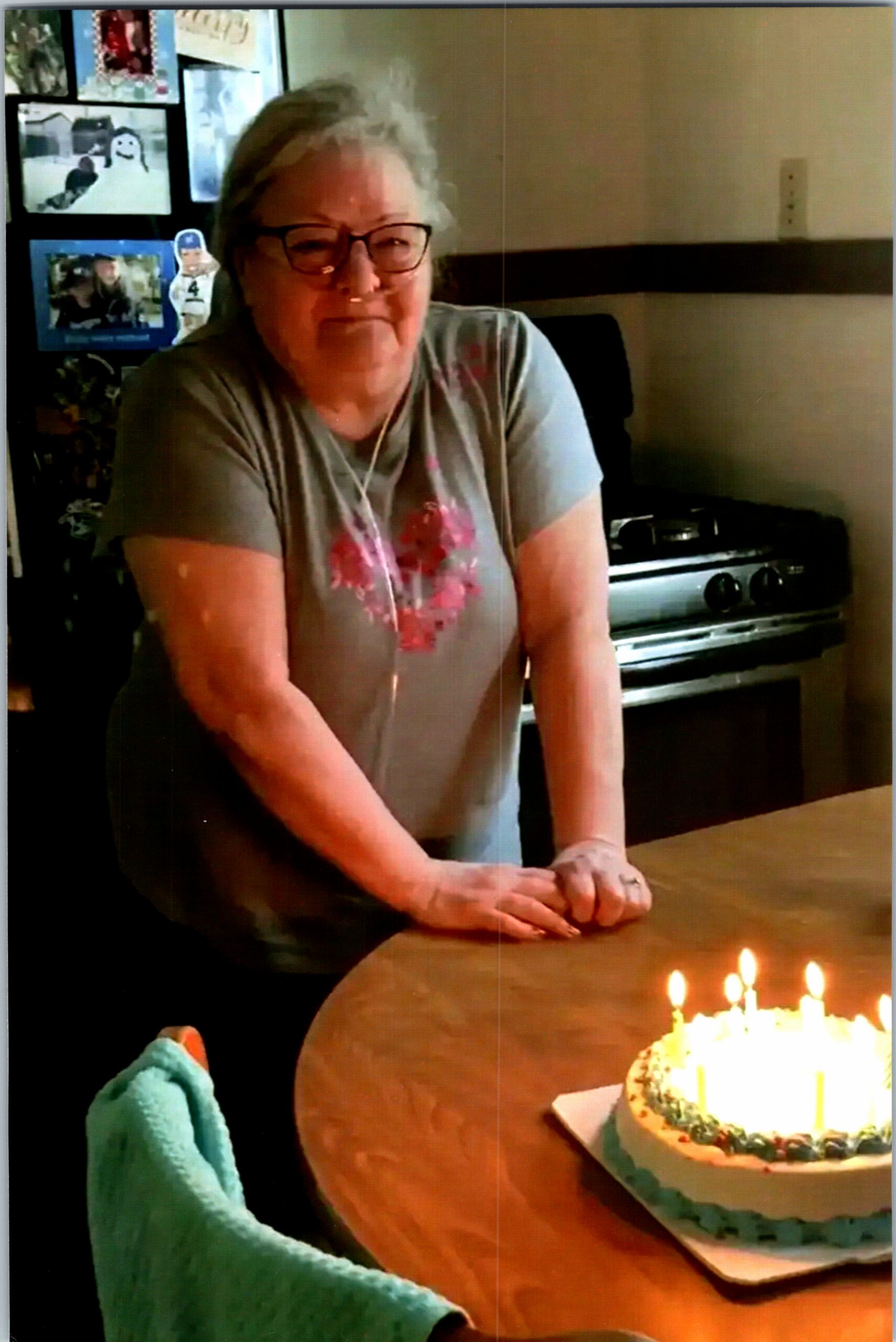 Older woman with glasses leaning on a kitchen table beside a birthday cake with lit candles.