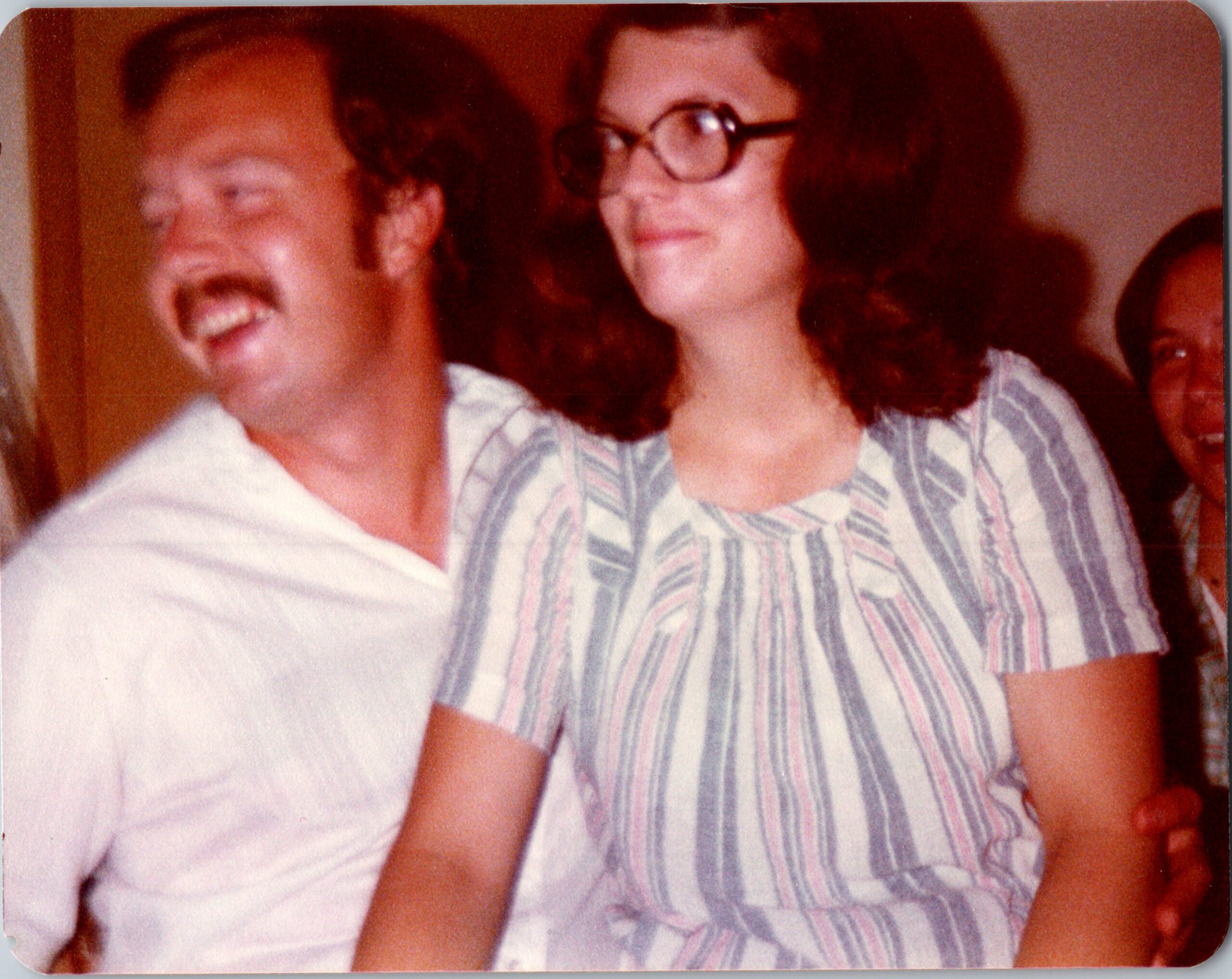 Two people smiling for a casual photo: a man with a mustache and white shirt on the left, and a woman with glasses wearing a striped blouse on the right.