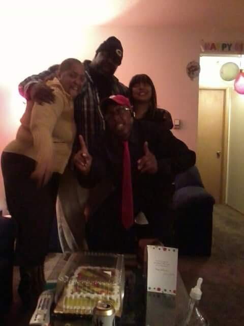 Five friends pose together in a living room birthday party, cake on the coffee table in front of them, balloons visible in the background.