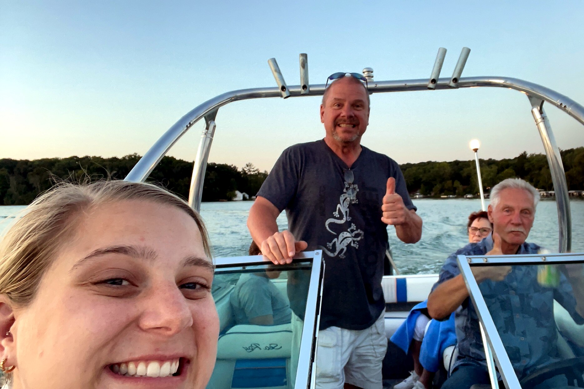 Close-up of a smiling woman taking a selfie on a boat, with a man behind her giving a thumbs-up and other passengers in the background on the water.
