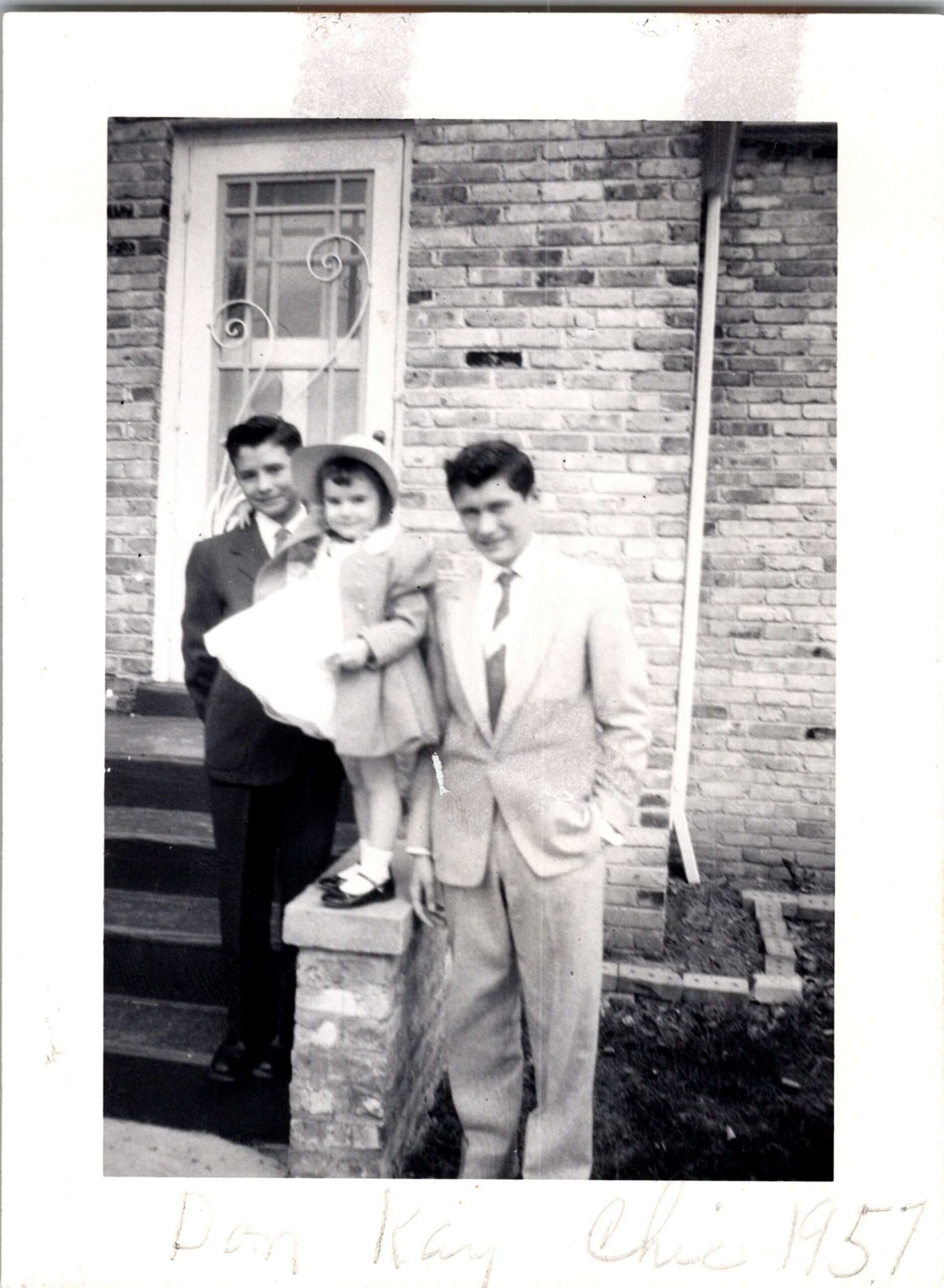 Three people in vintage clothing pose on the front steps of a brick house—two men in suits and a girl wearing a hat.