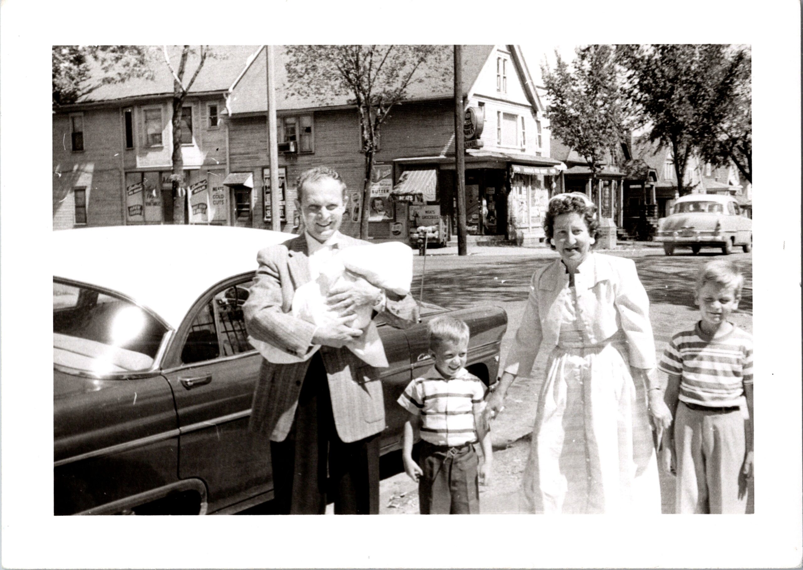 Man in a suit beside a vintage car, smiling, with two children and a woman posing on a sunlit city street with storefronts in the background.