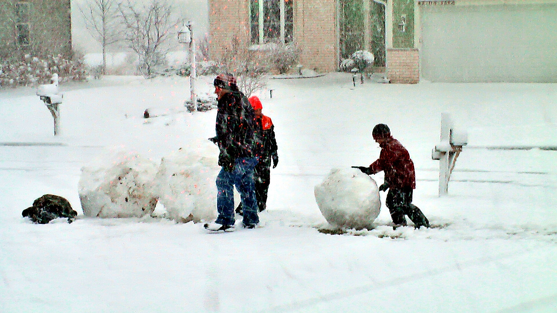 Two people and a child building a snowman in a snowy yard; a red-jacket child rolls a large snowball while an adult watches, with a dog resting nearby and houses in the background.