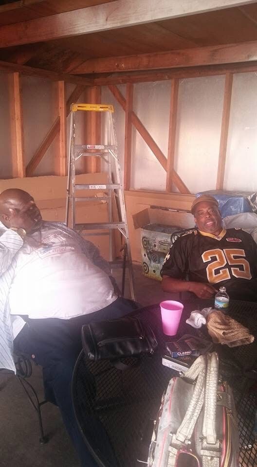 Two men sit at a table in a partially finished garage or attic; a step ladder stands in the background near wooden beams and plastic sheeting. 25 jersey on one man.
