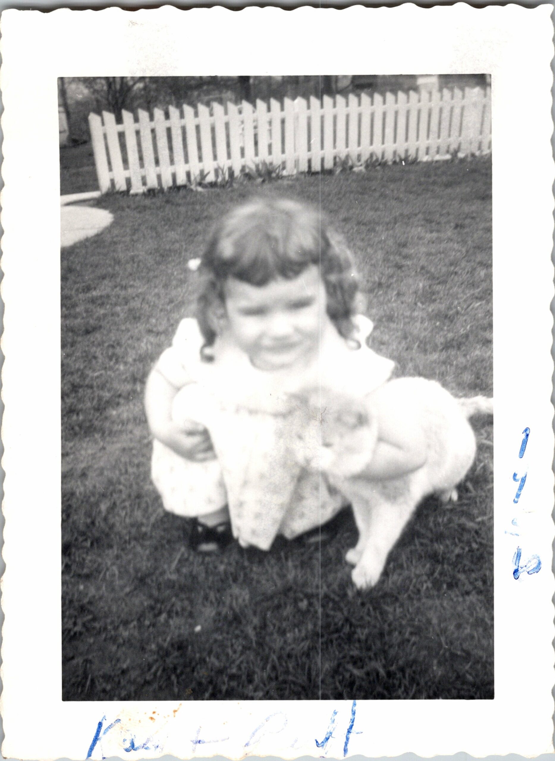 Blurry black-and-white photo of a young girl with pigtails wearing a light dress, squatting on grass in front of a white picket fence.