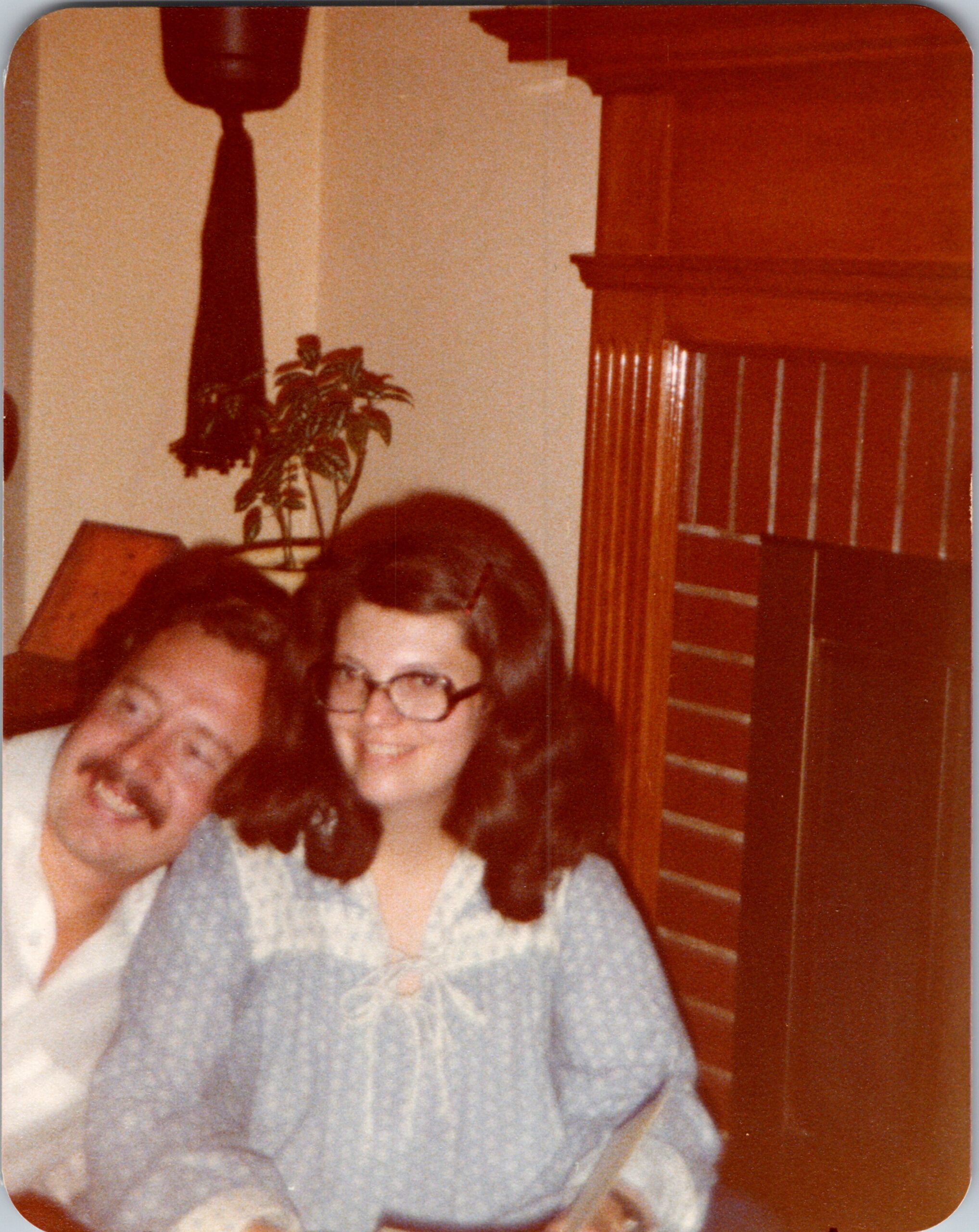 Smiling man and woman pose for a photo in a living room with a wooden cabinet and a potted plant.