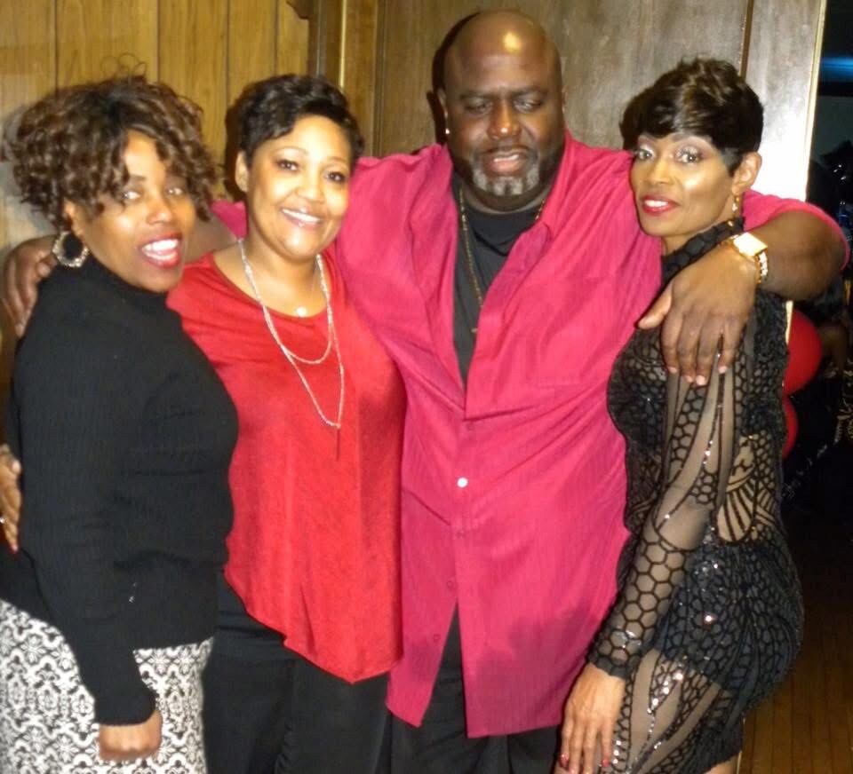 Four adults pose together with arms around each other at a social gathering, smiling for the camera in a warm group photo. The man in the center wears a pink shirt, flanked by two women in black outfits and another woman in a red top.