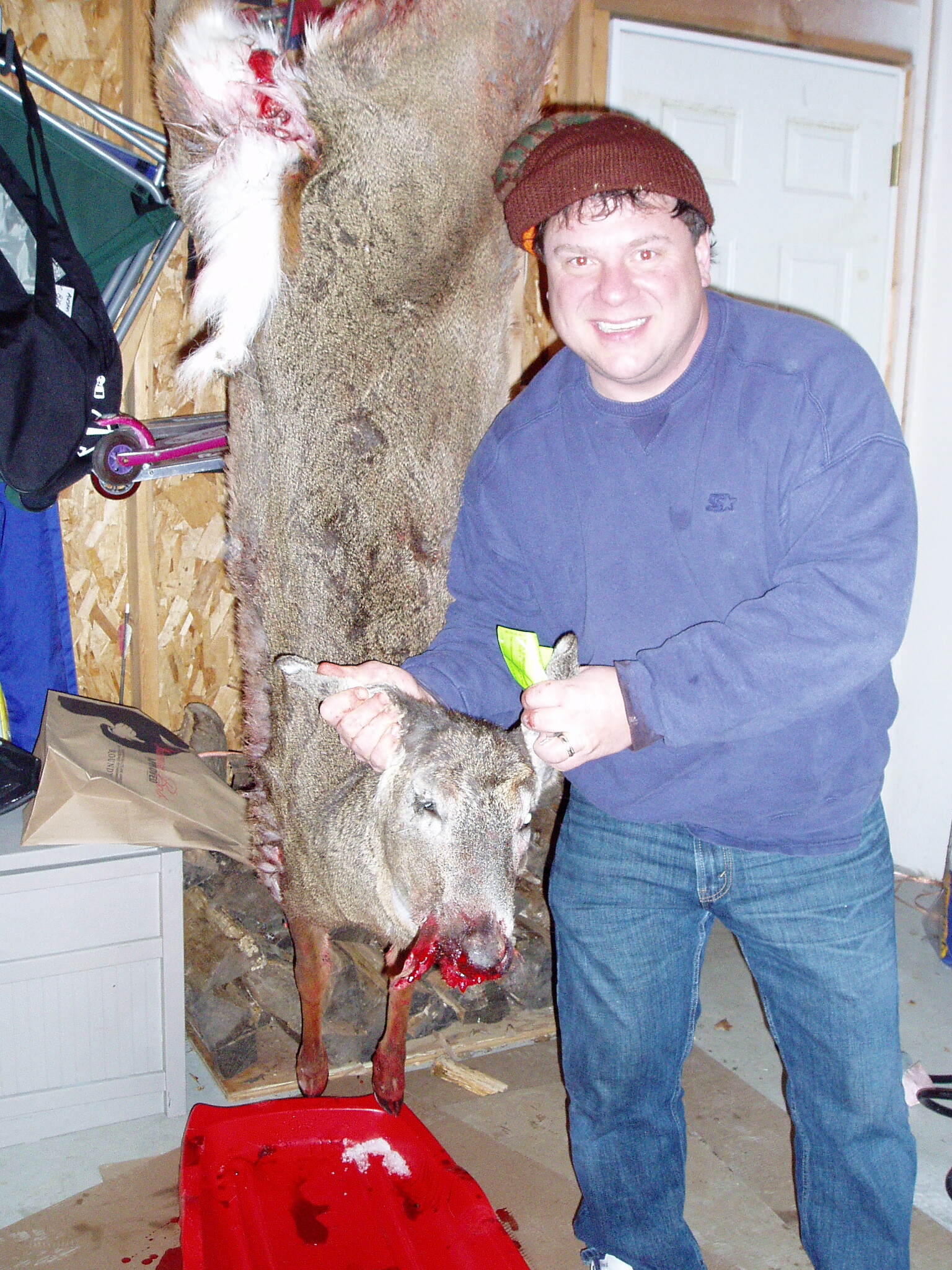 Man in a blue sweatshirt and brown knit cap posing with a dead deer hanging in a garage; blood on the floor nearby.