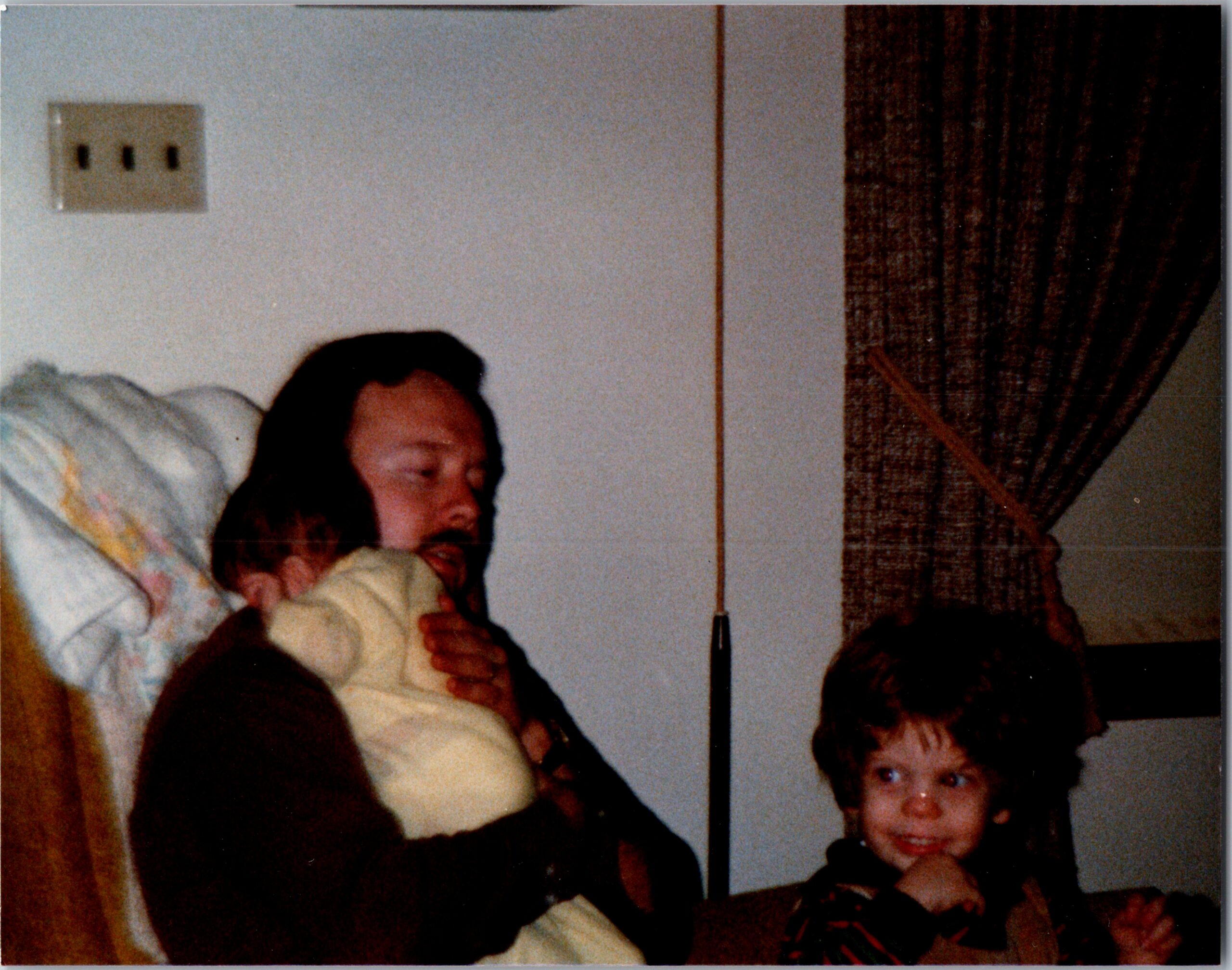Woman cradling a sleeping baby on a sofa, with a child in the foreground and dark curtains to the right.
