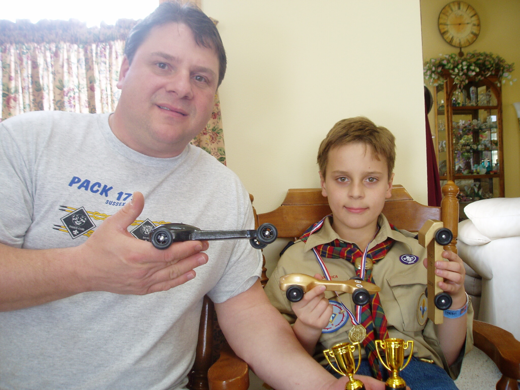 Two smiling people posing indoors, the man holding a black toy car while a boy in a scout uniform shows more toy cars and trophies on his lap.