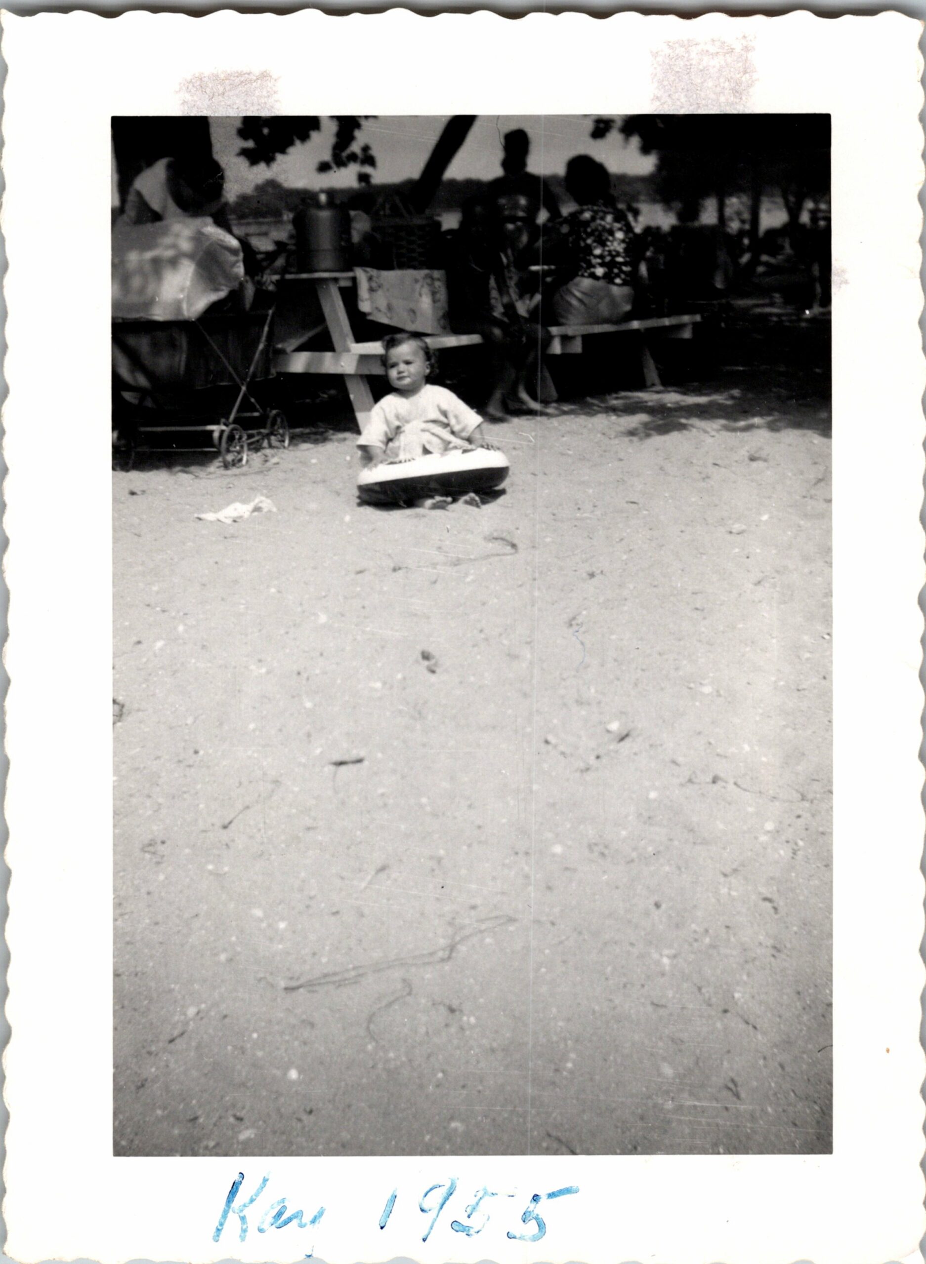 Toddler sitting in a round toy on a sandy park ground, with people and a stroller in the background.