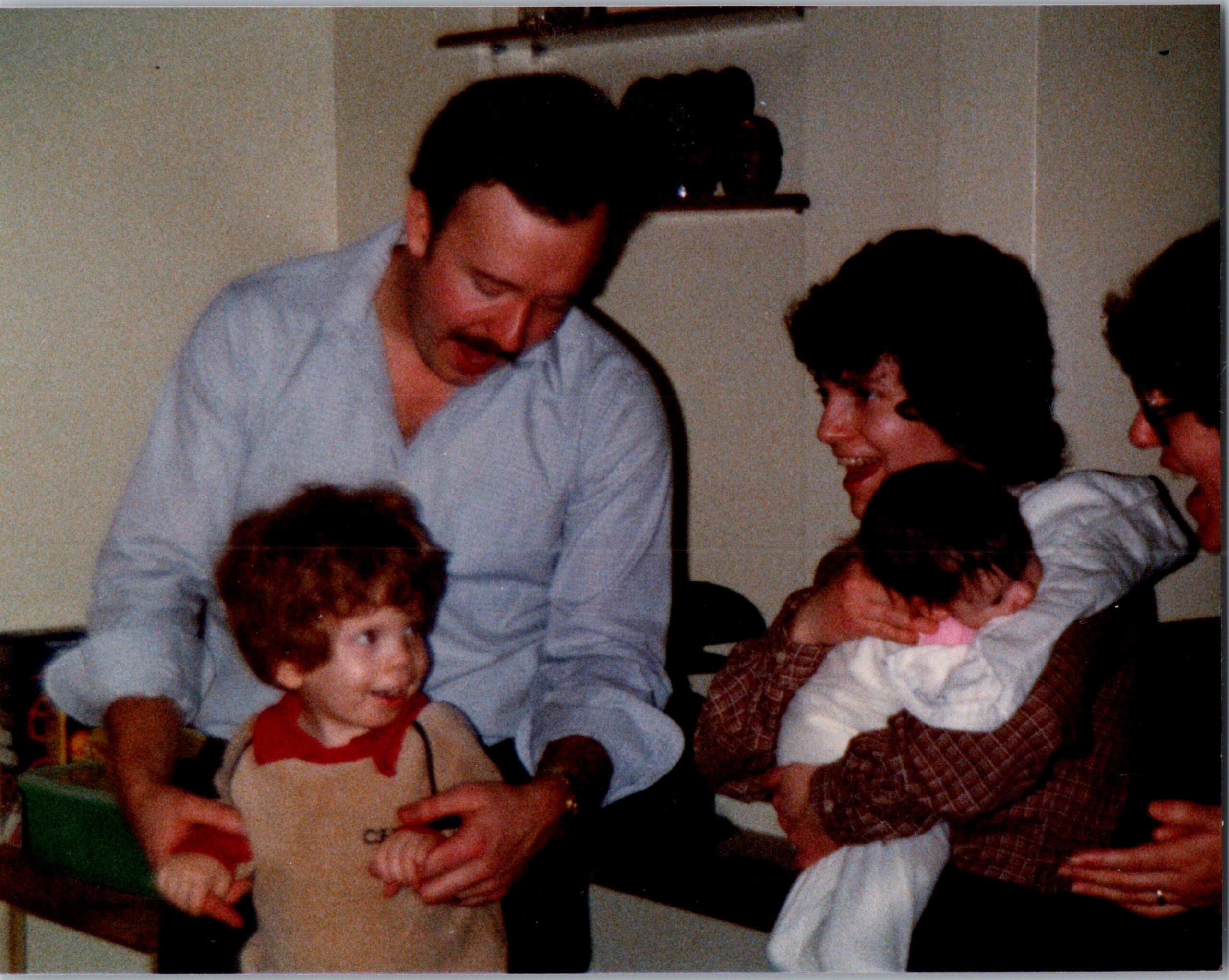 Man in a light blue shirt leans over, smiling, while a little girl holds a large book and others look on indoors.