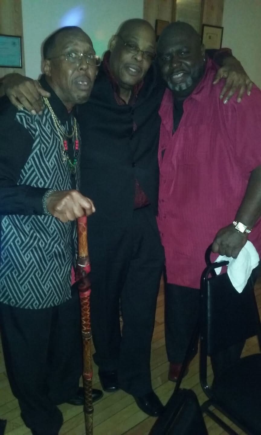 Three men pose together, smiling with arms around each other at a social event; one holds a decorated cane, another uses a walker nearby.