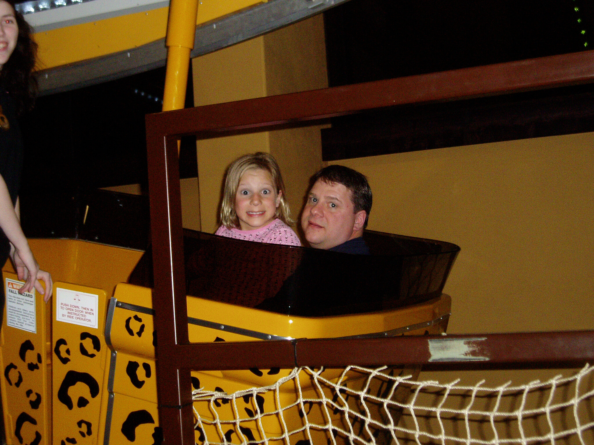 Two riders, a man and a young girl, sit in a yellow amusement-ride car and smile at the camera.