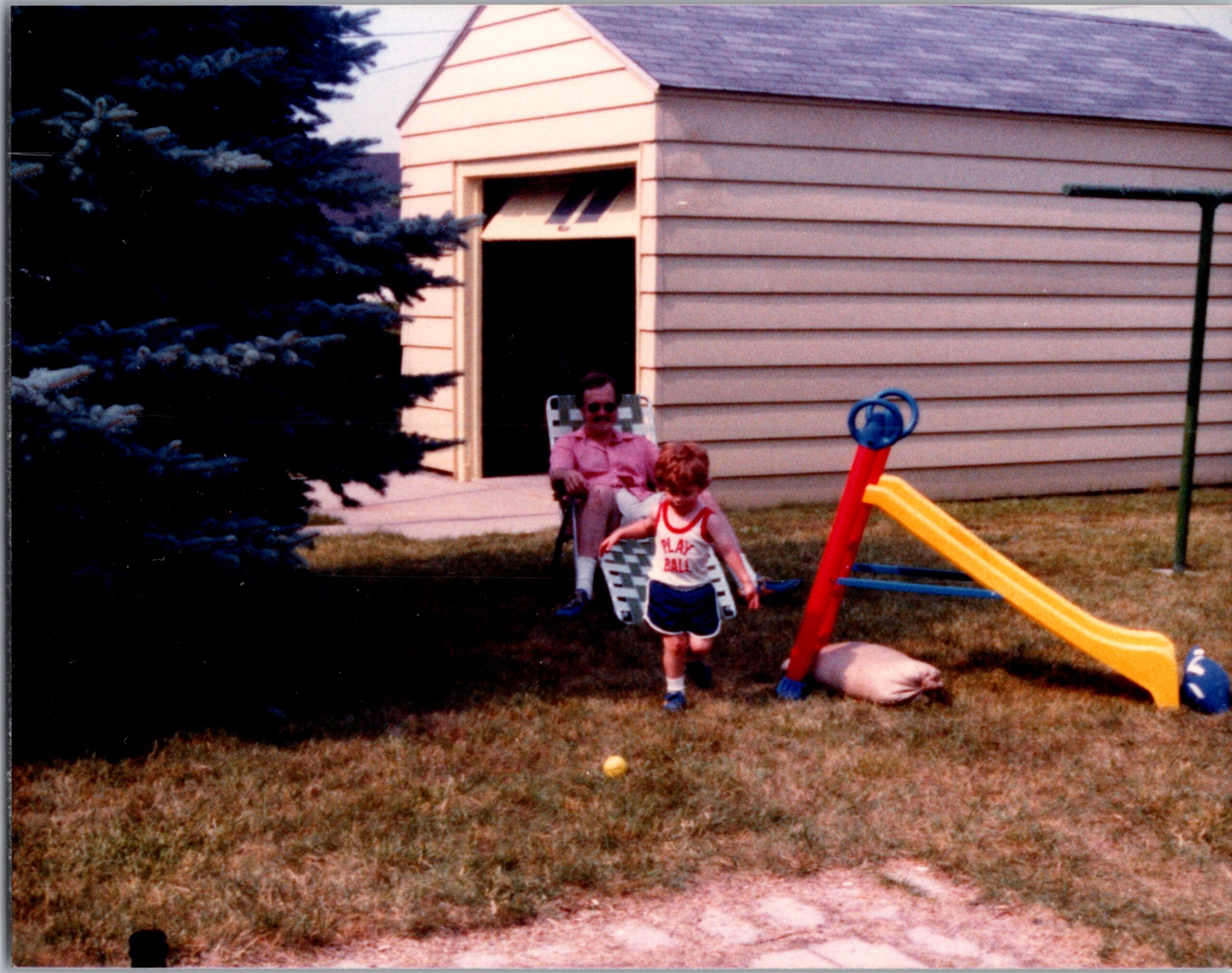 Backyard scene with a yellow and red plastic slide, two children playing near a reclining chair and a beige house with horizontal siding, and a dark evergreen tree on the left.