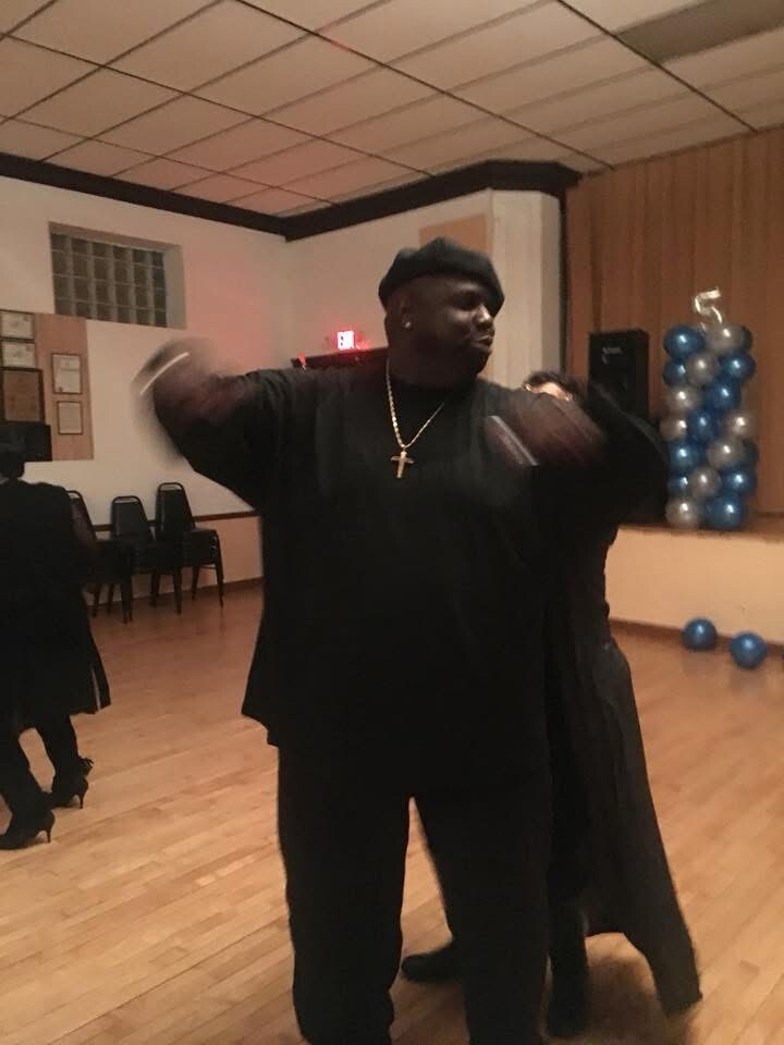 A man in a black outfit and cross necklace dances with arms raised in a community hall, blue balloon decorations in the background.