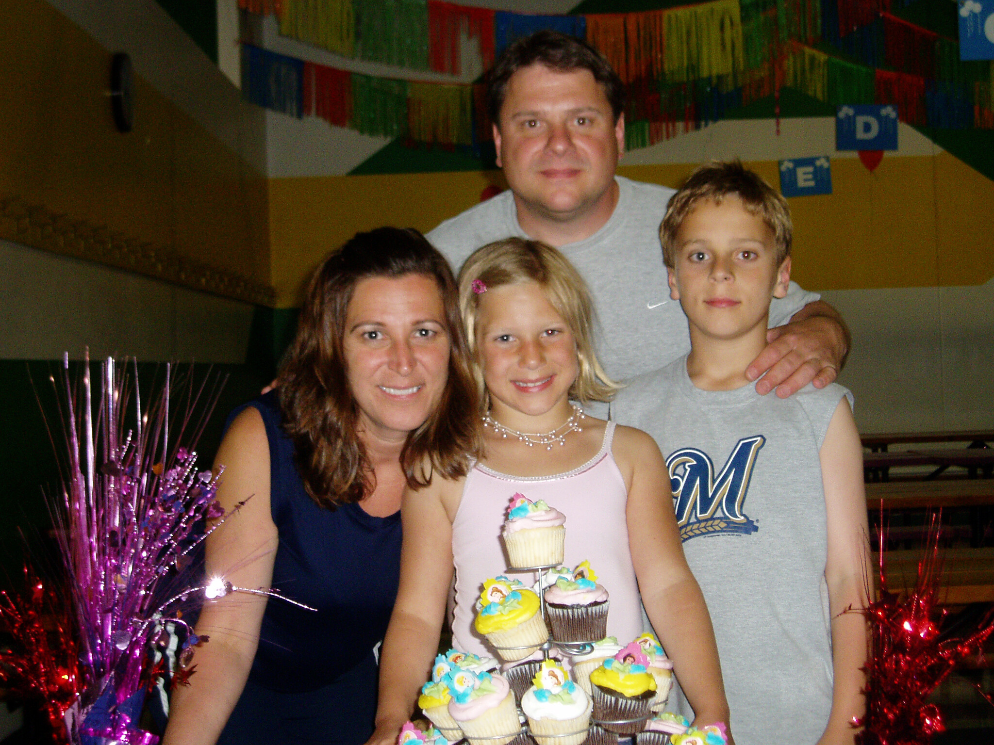 Four people smile for a photo at a party, with a tall cupcake tower centerpiece and pink tinsel decorations nearby.