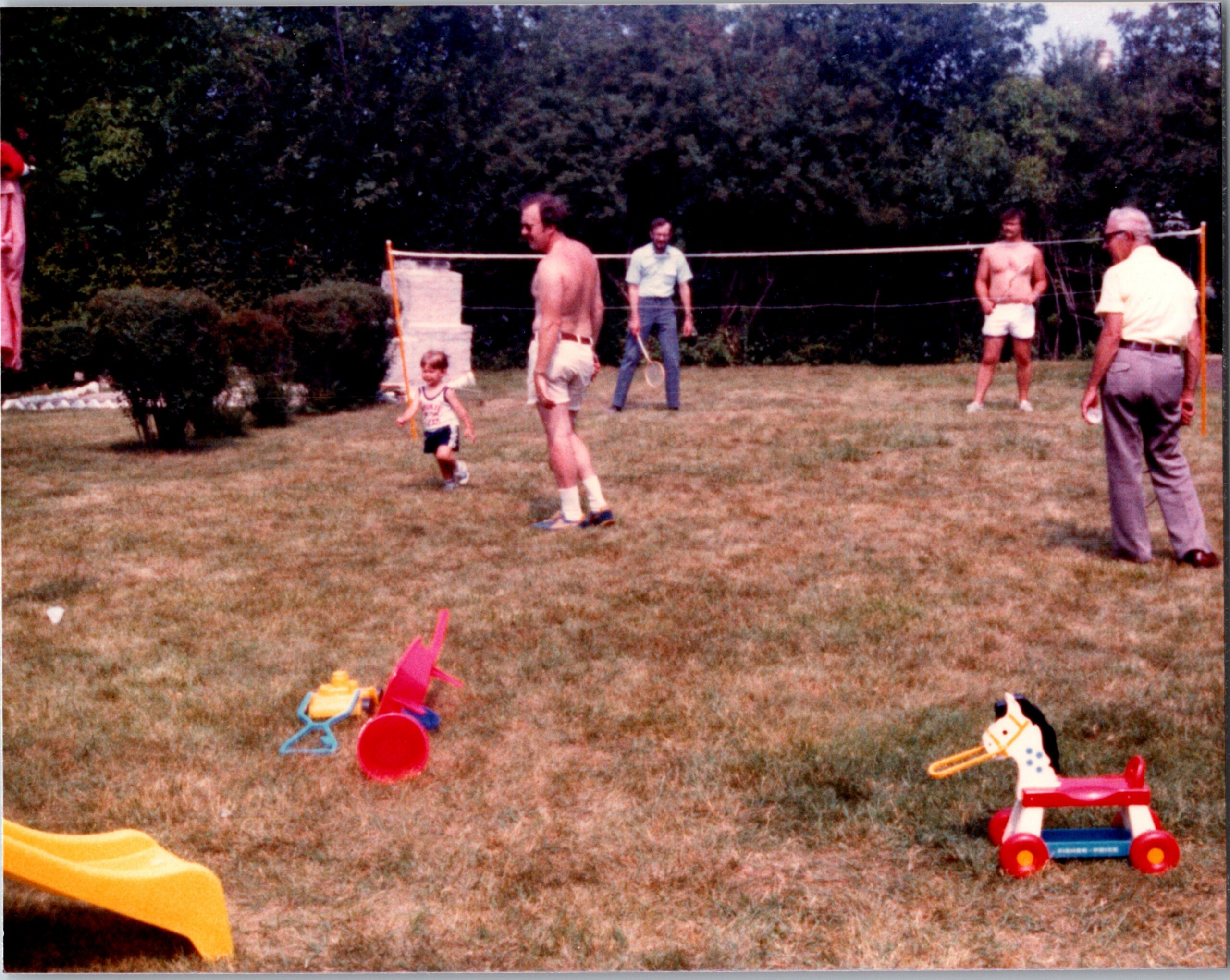 People playing in a backyard with a low badminton-style net, a child runs with a toy bat while adults supervise nearby, toys scattered on the grass.
