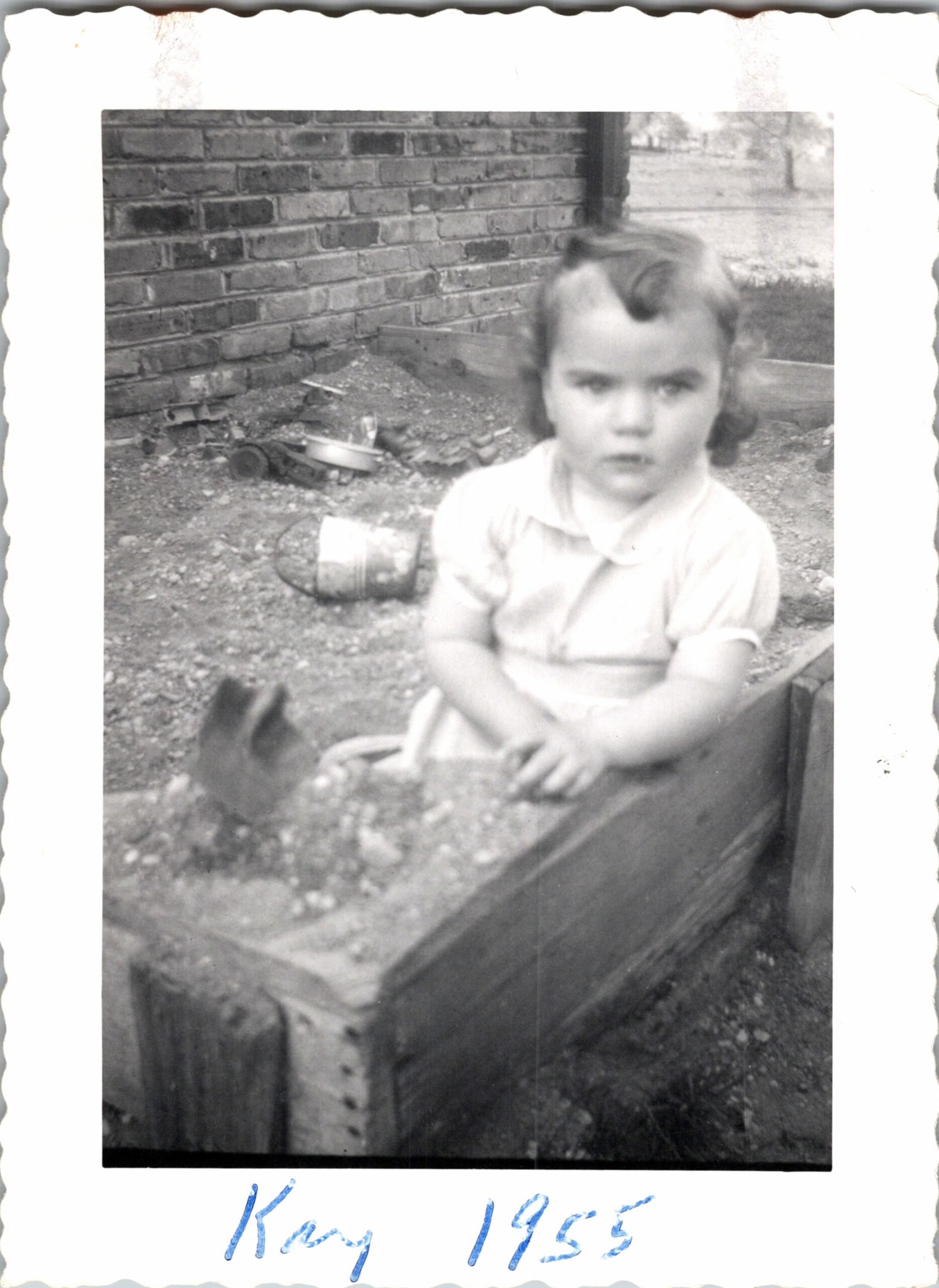 Black-and-white photo of a young child sitting outdoors beside a brick wall, with scattered gravel and a toy on the ground, looking toward the camera.