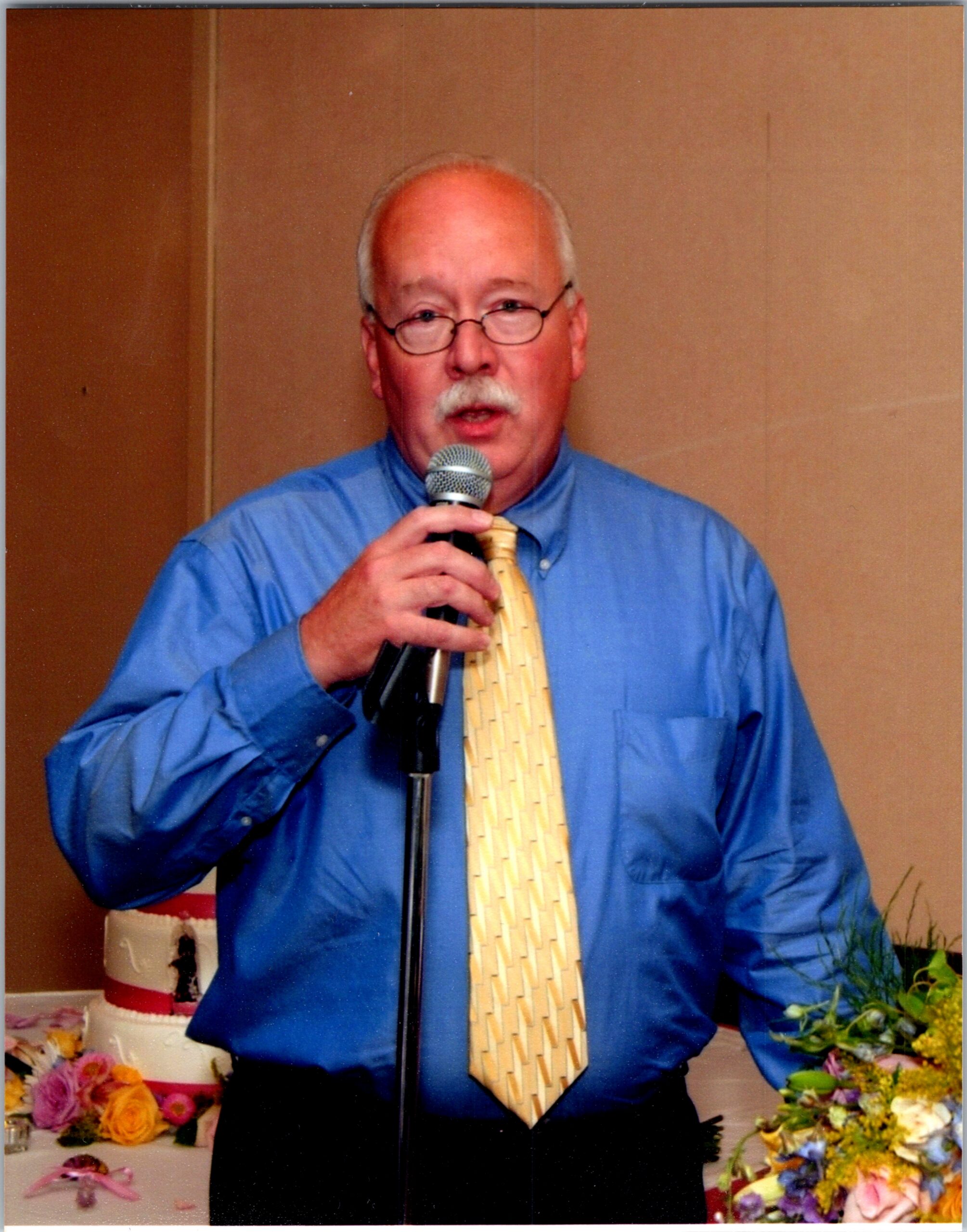 Older man with glasses speaking into a microphone, wearing a blue shirt and yellow tie, with colorful flowers on a table in front.