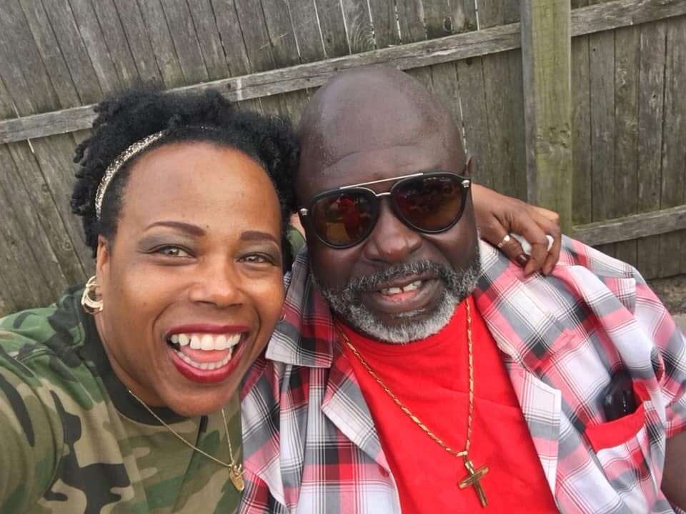 Woman and man take a cheerful selfie outdoors, smiling at the camera in front of a wooden fence.
