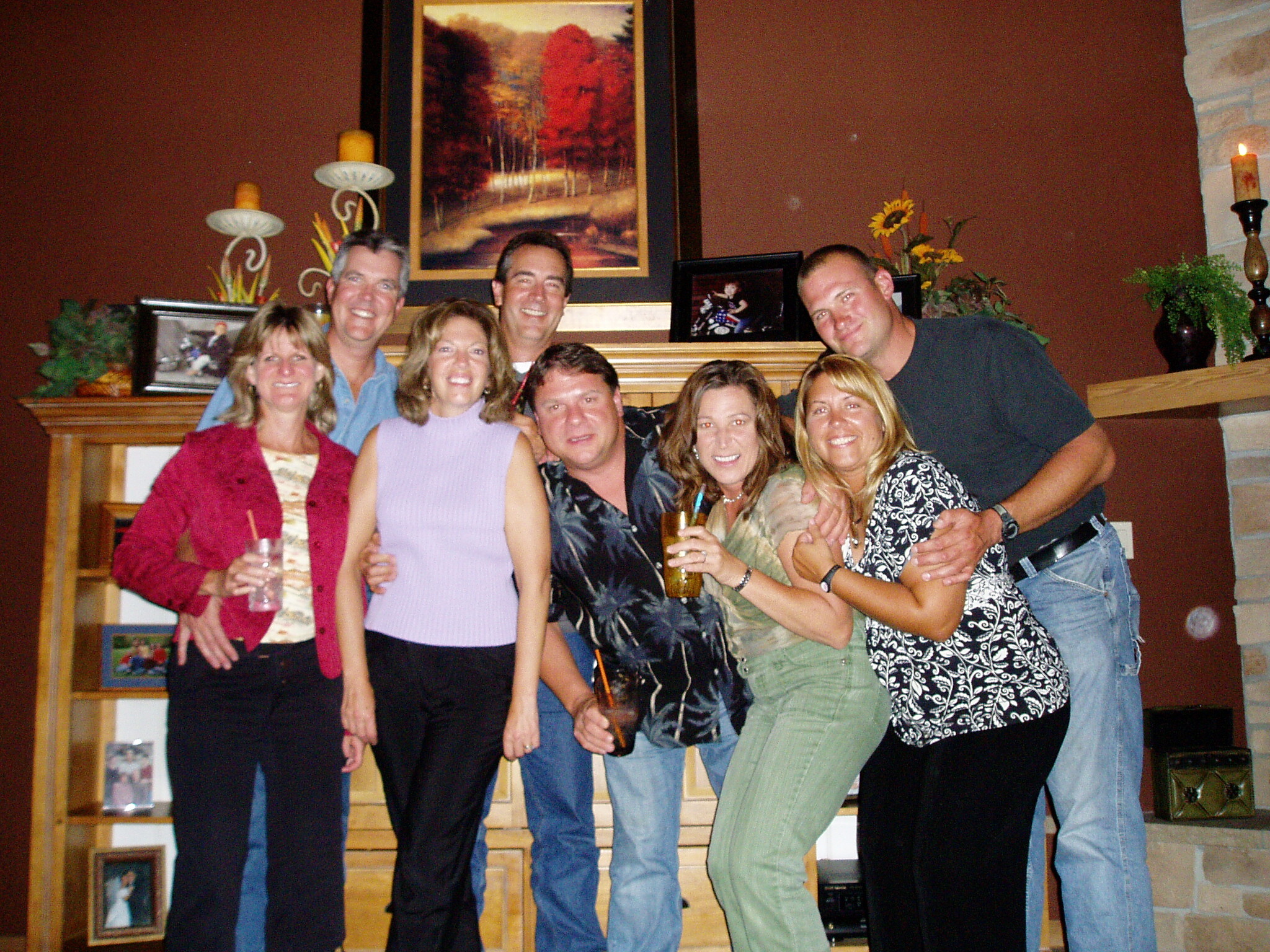 Group of eight adults posing and smiling in a living room, some holding drinks in front of a fireplace and decorative art.