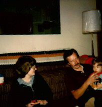 Three people sitting on a dark couch in a living room; a woman on the left looks toward a man on the right holding a small child.